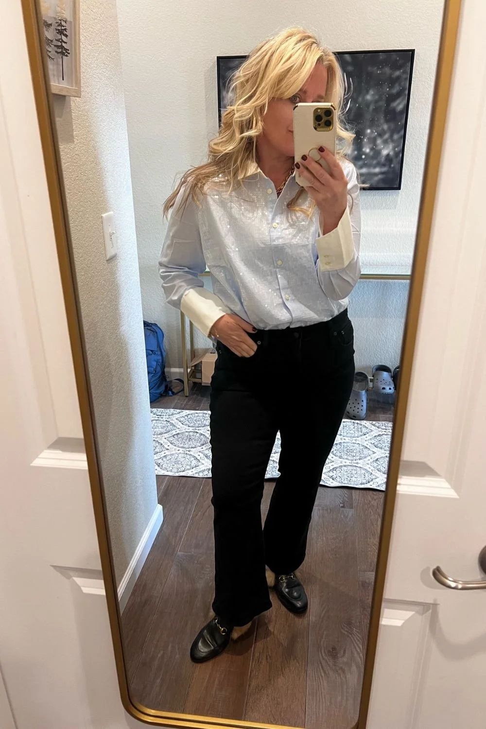 A woman with blonde curly hair taking a mirror selfie in a room. She is wearing a shiny white button-up shirt, black pants, and black shoes. There is a television on the wall behind her, and a small table with items on it, including a blue backpack on the floor. The room has a patterned rug and a light-colored wall.