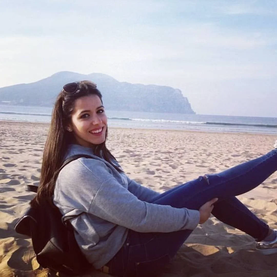 A woman sitting on a sandy beach, smiling, with a mountain and ocean in the background.