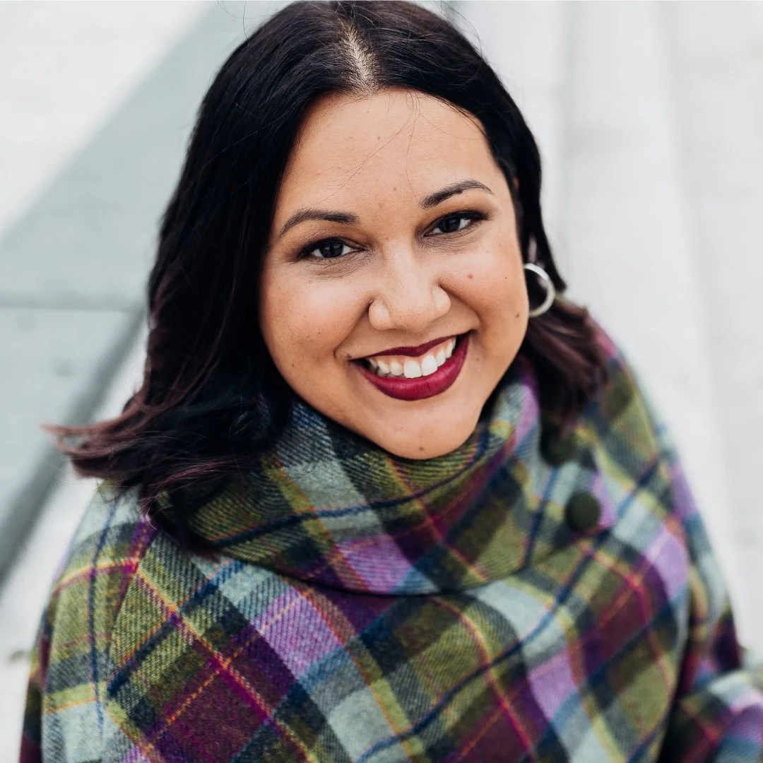 Close-up of a smiling woman with dark hair, wearing earrings and a colorful plaid top, outdoors against a light background.