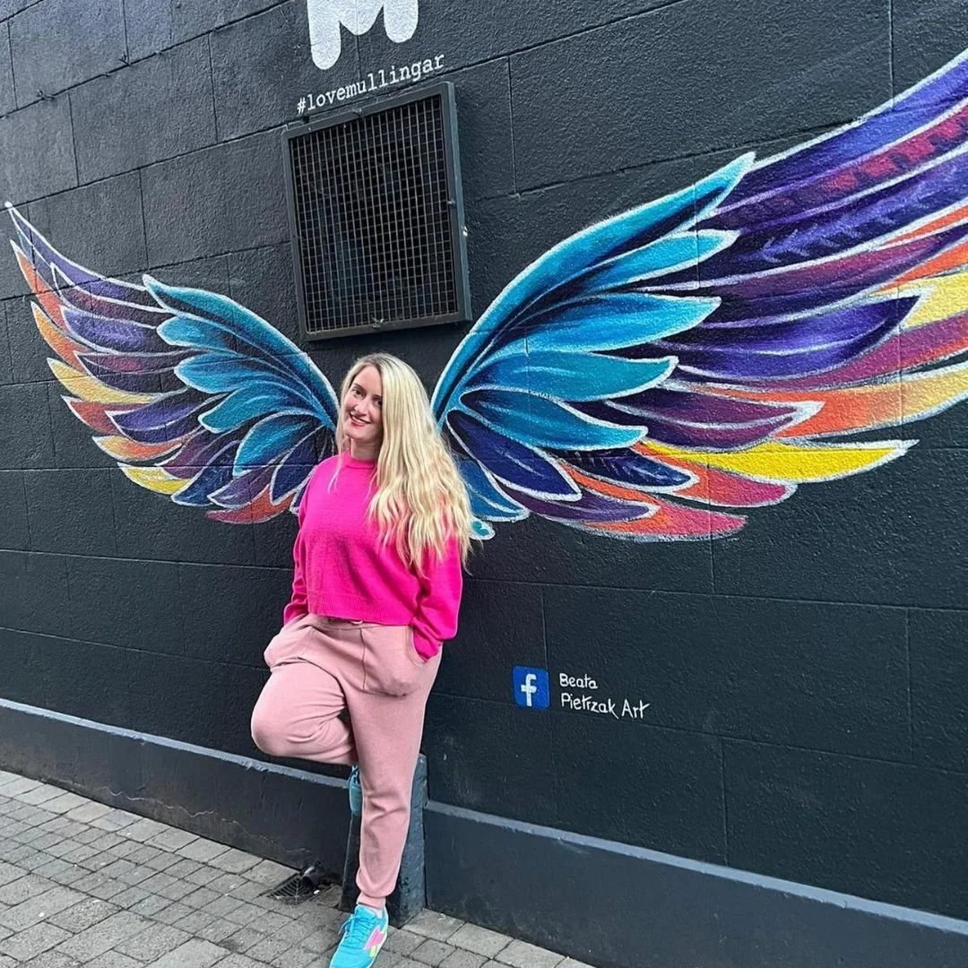 A woman in pink clothing standing in front of a mural of colorful wings on a black wall, with a Facebook logo and text that says Beata Pietzak Art.