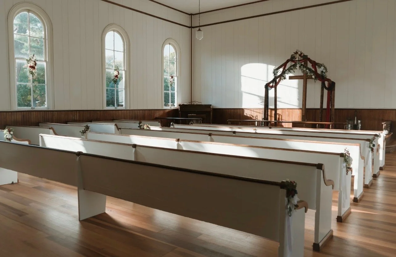Empty church with white pews decorated for a wedding, floral arrangements on the ends, and a wedding arch draped with greenery and red ribbons at the front, with three tall arched windows letting in natural light.