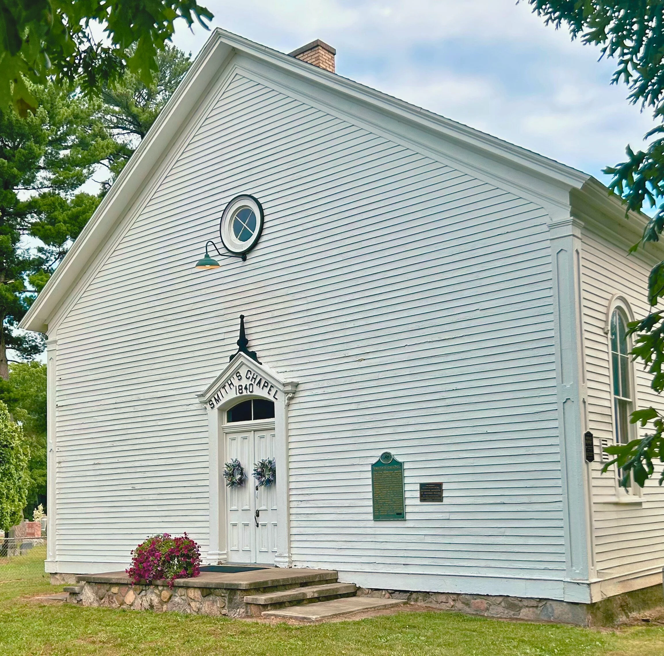 White historic chapel with a gabled roof, double doors decorated with floral wreaths, and signs indicating it is Smith's Chapel, built in 1840. There are steps leading up to the entrance, with a flower bed of pink flowers in front. A small circular w