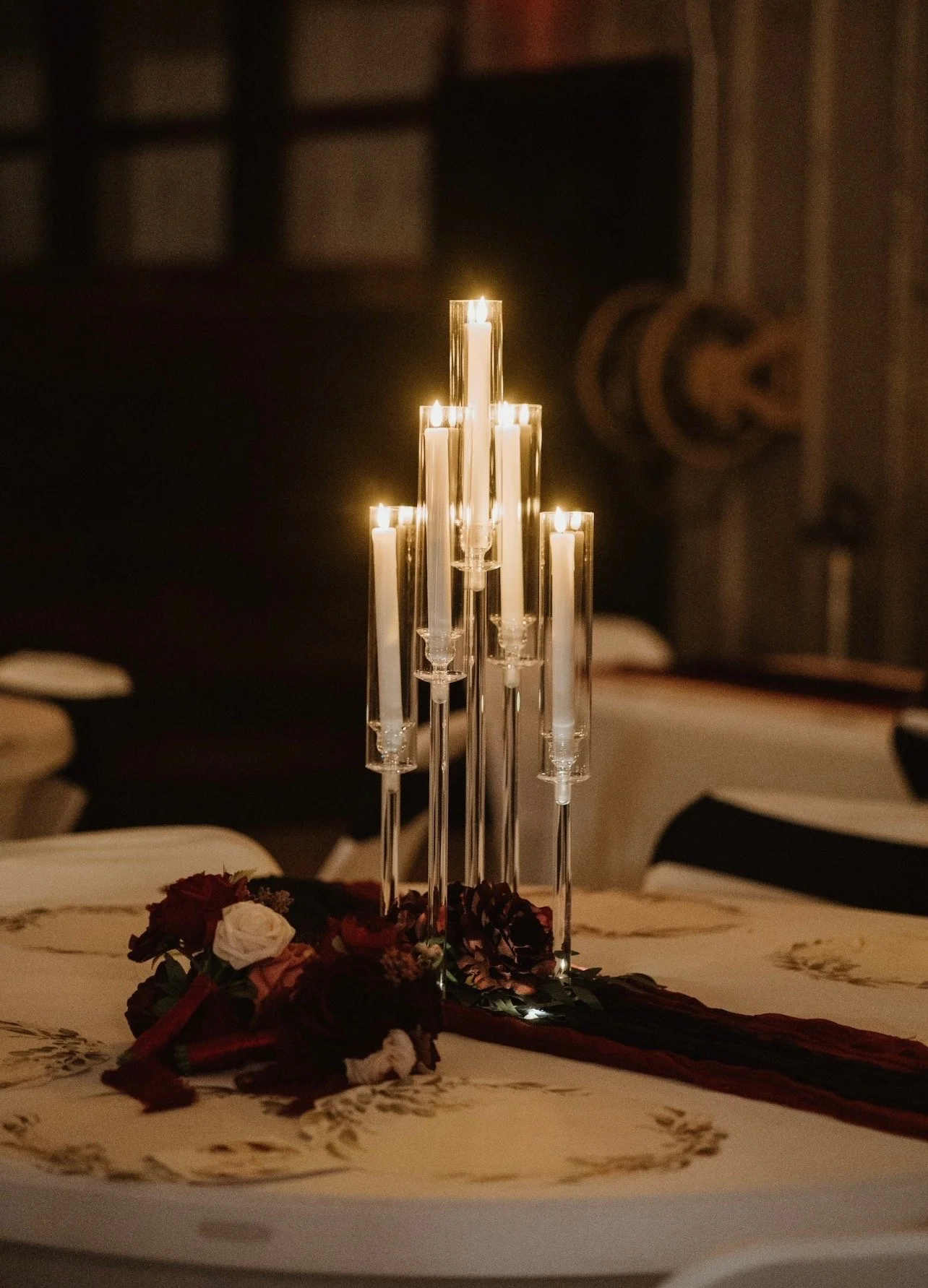 Table centerpiece with tall, lit, white candles in clear glass holders and a bouquet of dark red and white flowers on a white tablecloth in a dimly-lit room.