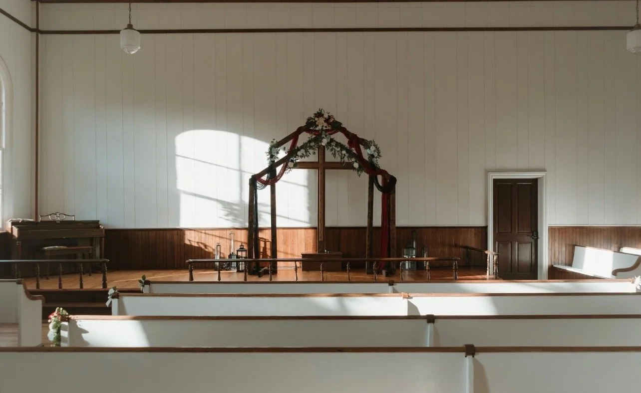 Empty church interior with a decorated wooden cross on stage, floral arrangements, lanterns, and white pews.