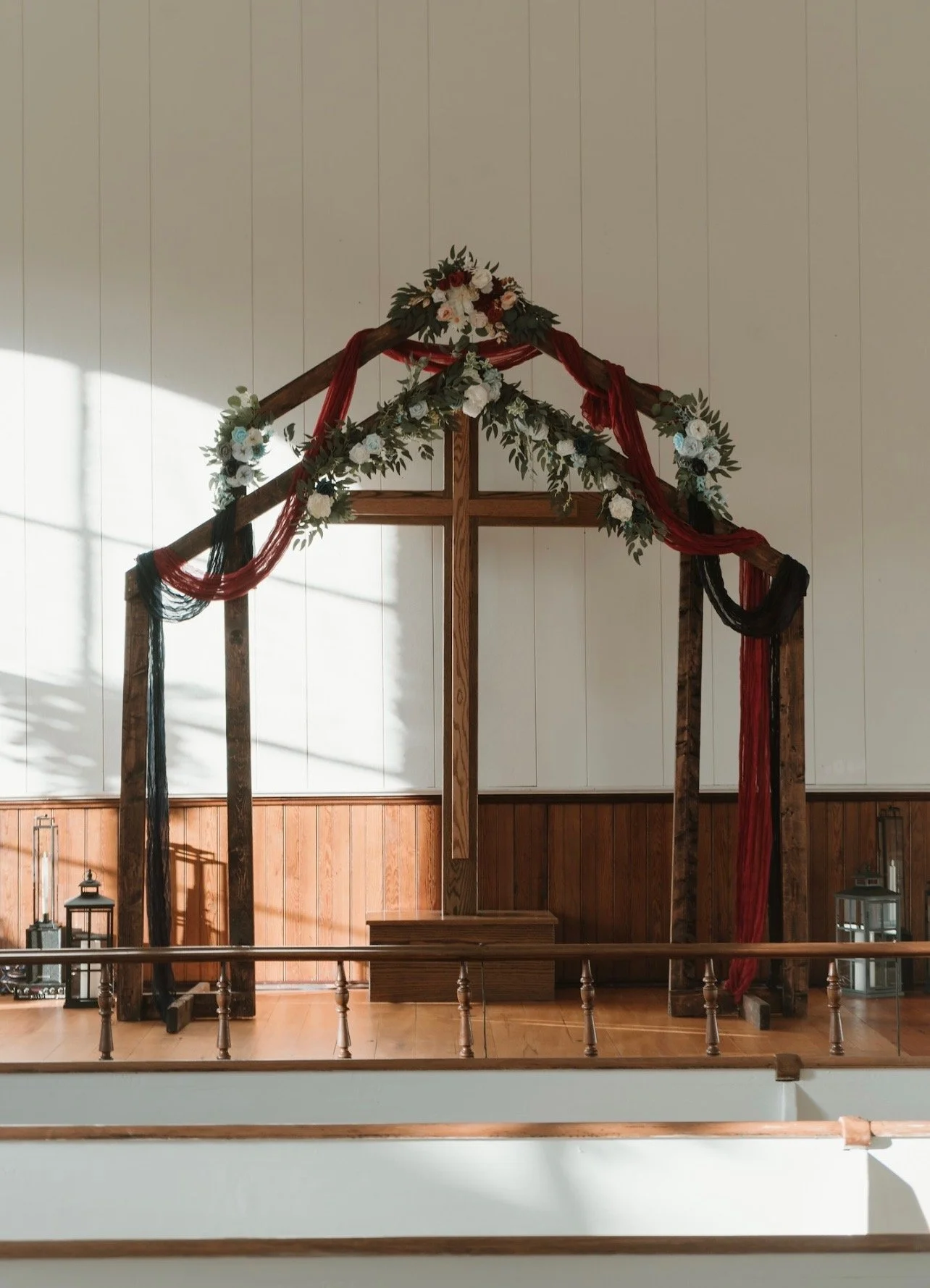 A church altar with a wooden cross decorated with red, black, and white fabric and white flowers with green foliage, set against a white wall with wooden paneling.