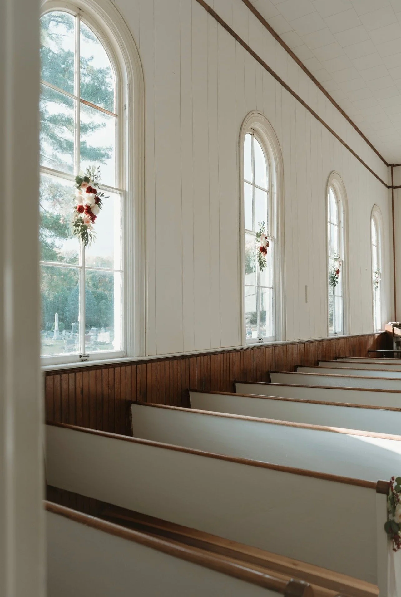 Interior of a church with tall arched windows decorated with flowers, wooden pews, and white walls.