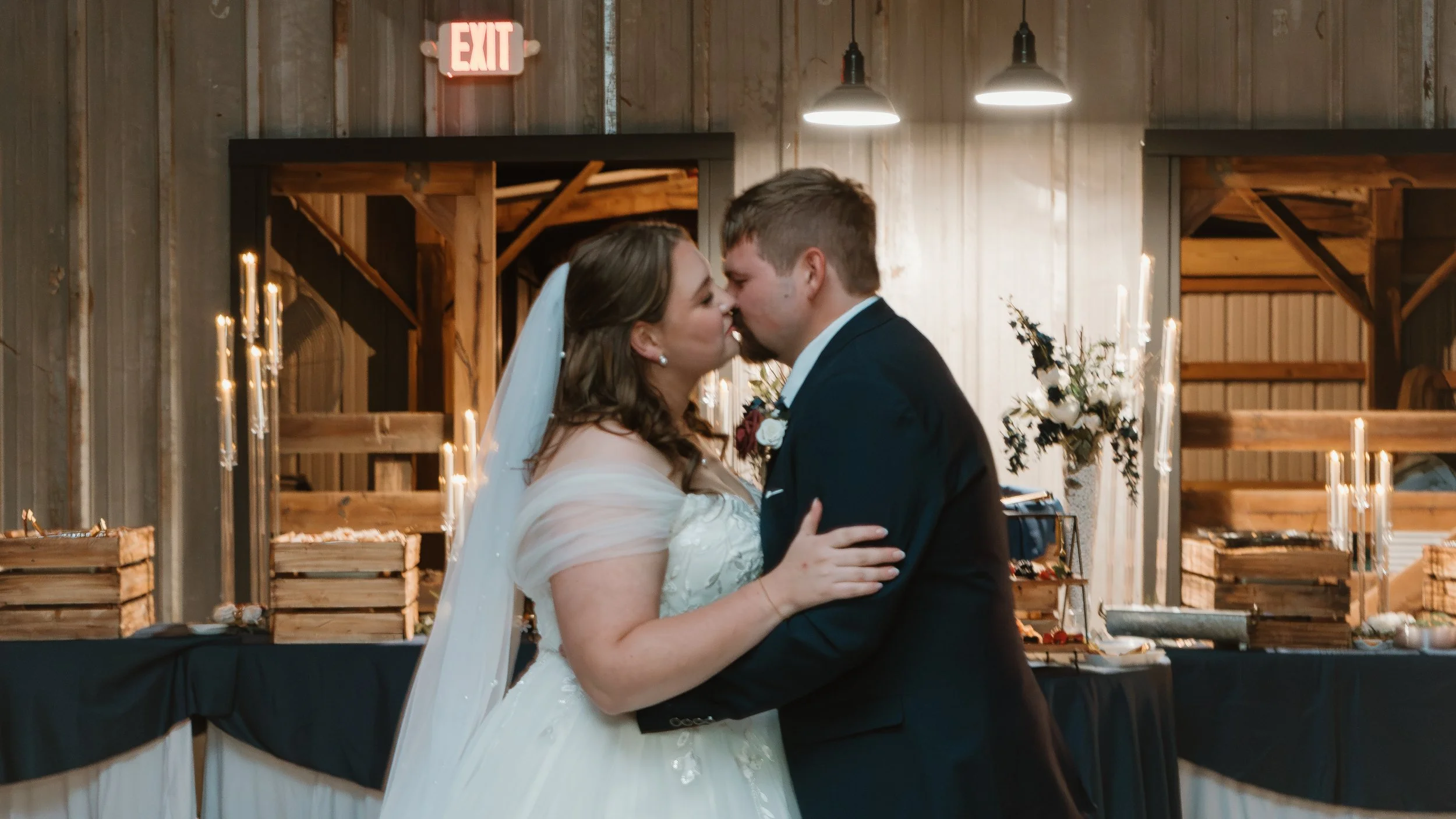 A bride and groom share a kiss during their wedding reception in a rustic barn, with candles and flowers in the background.