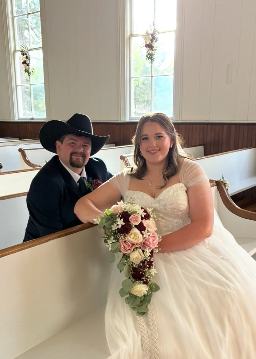 A bride and groom sitting inside a church with white walls and wooden pews, with large windows behind them, holding a bouquet of flowers.