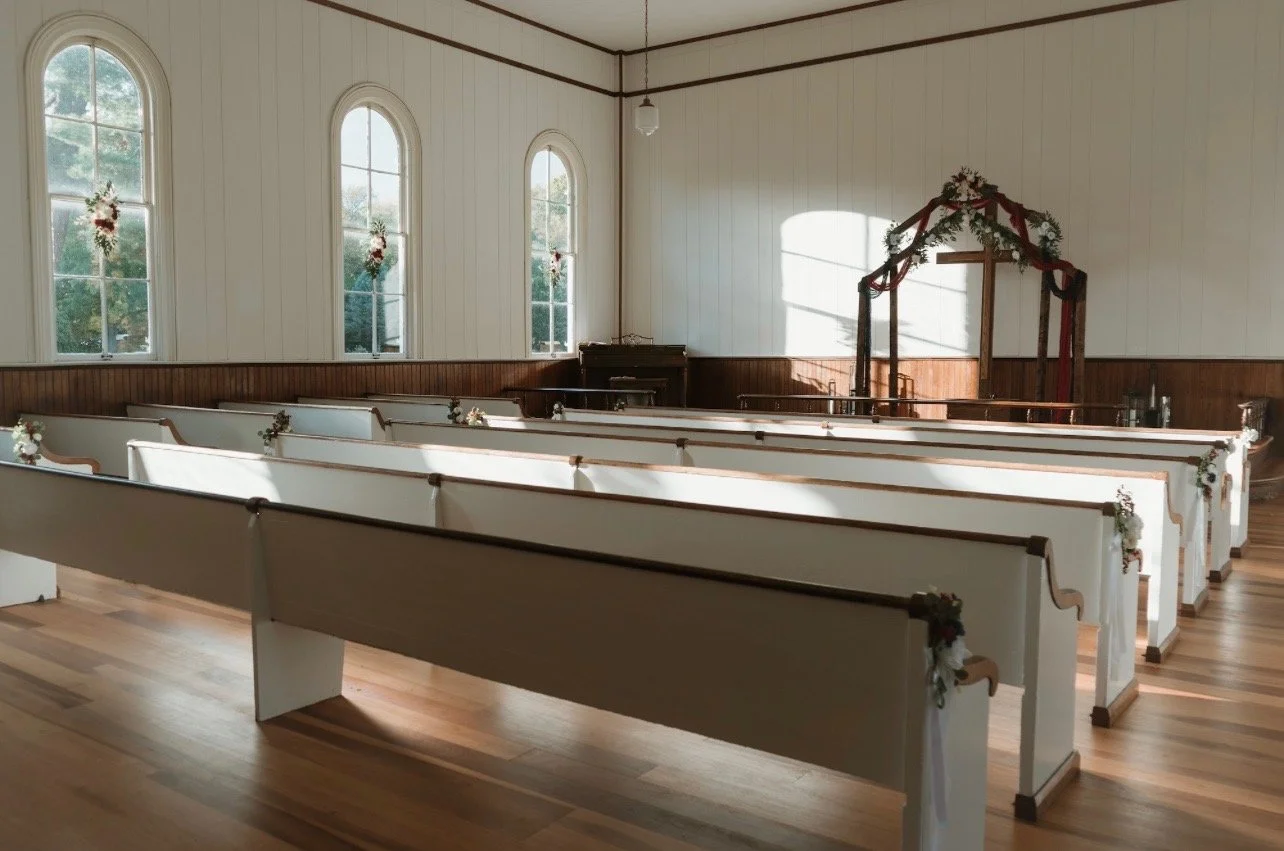 Interior of a small, decorated church with white pews, floral arrangements on the ends, large arched windows, and an altar with floral and ribbon decorations.