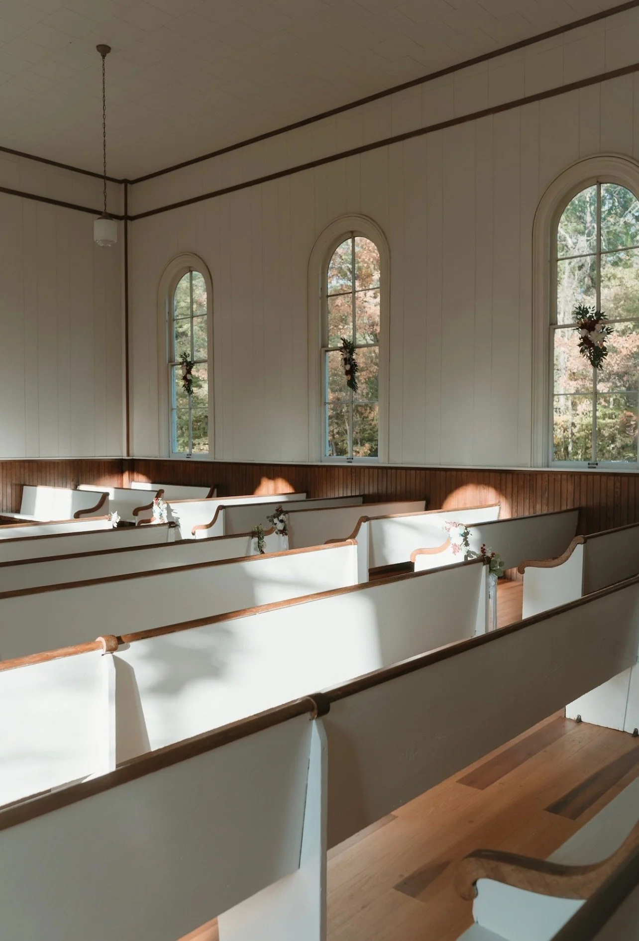 Interior of a small church or chapel with white pews decorated with small floral arrangements, tall arched windows with small wreaths, and sunlight casting shadows inside.