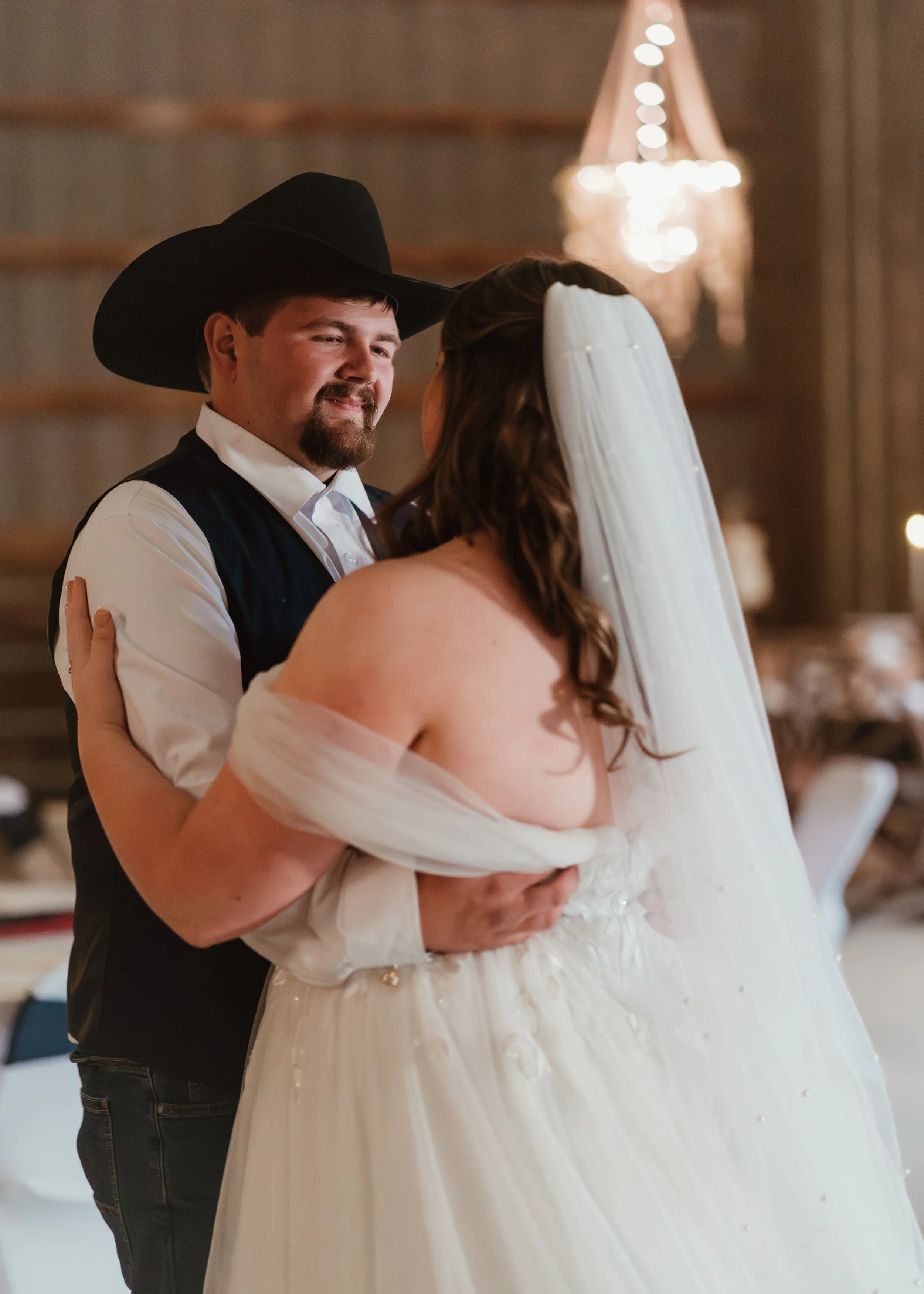 A couple sharing a dance at their wedding, the groom wearing a cowboy hat and the bride in a wedding dress with a veil.