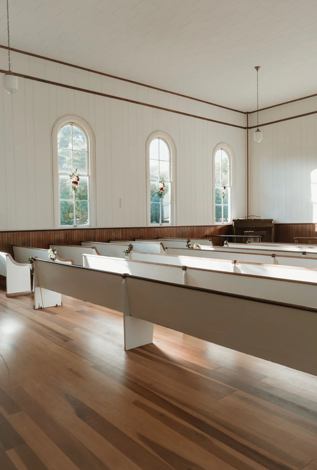 Interior of a small church with wooden pews decorated with small floral arrangements, three tall arched windows with floral decorations, and a wooden piano near the front.