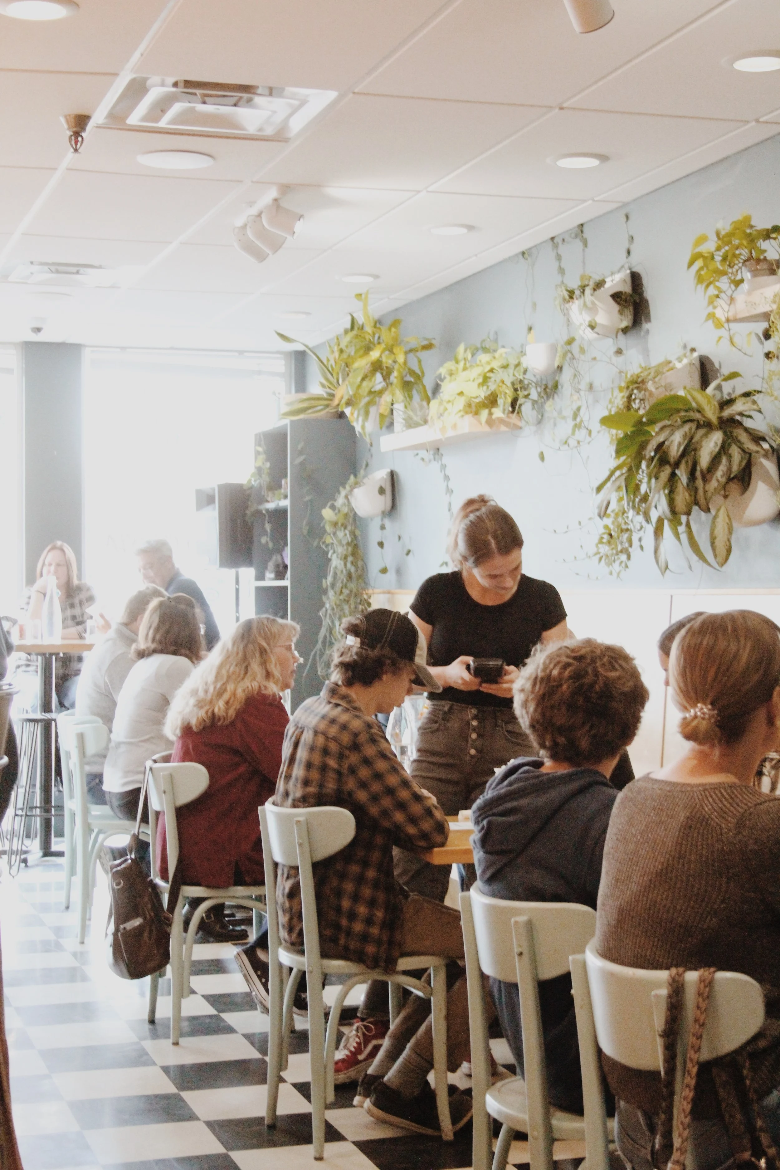 People sitting at tables and a woman standing in a cafe with potted plants on the wall.