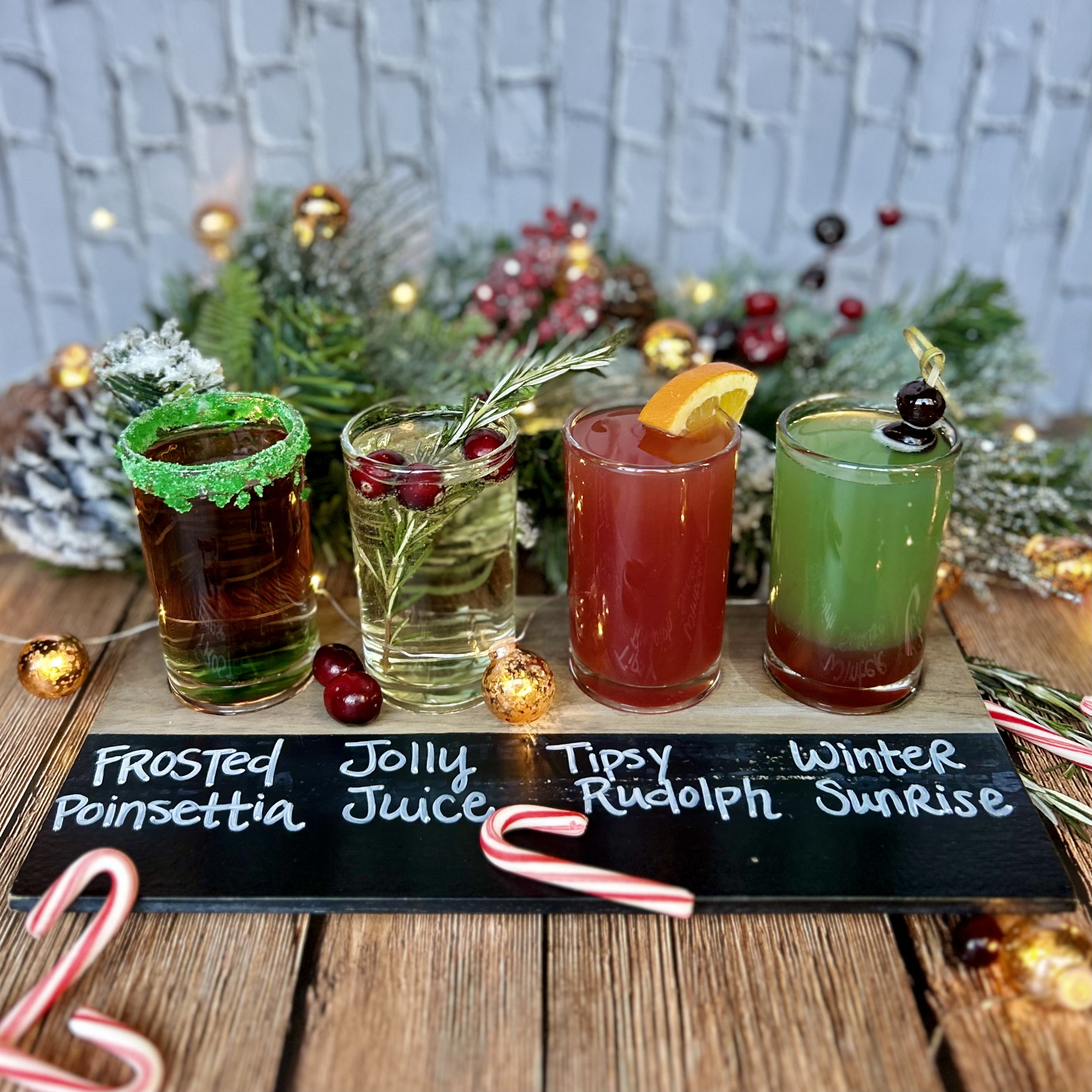 Four colorful holiday cocktails on a wooden table with a chalkboard label that reads 'Frosted Poinsettia', 'Jolly Juice', 'Tipsy Rudolph', and 'Winter Sunrise', decorated with seasonal ornaments and greenery in the background.