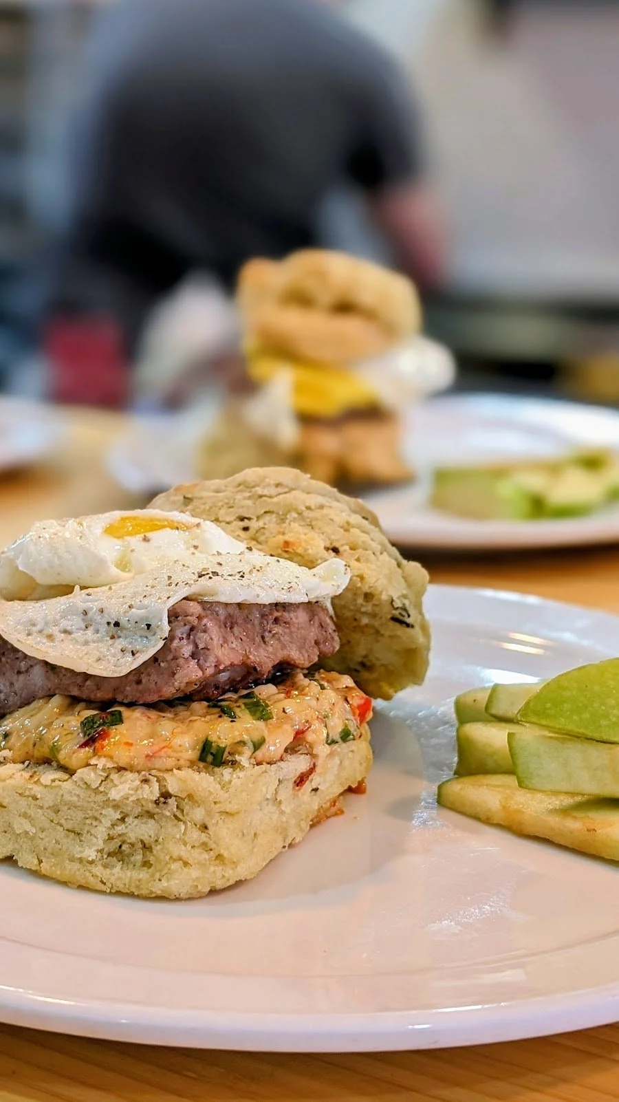 Plate with a breakfast sandwich topped with a fried egg, a biscuit, and a biscuit with a breakfast sausage patty, served with sliced green apples.