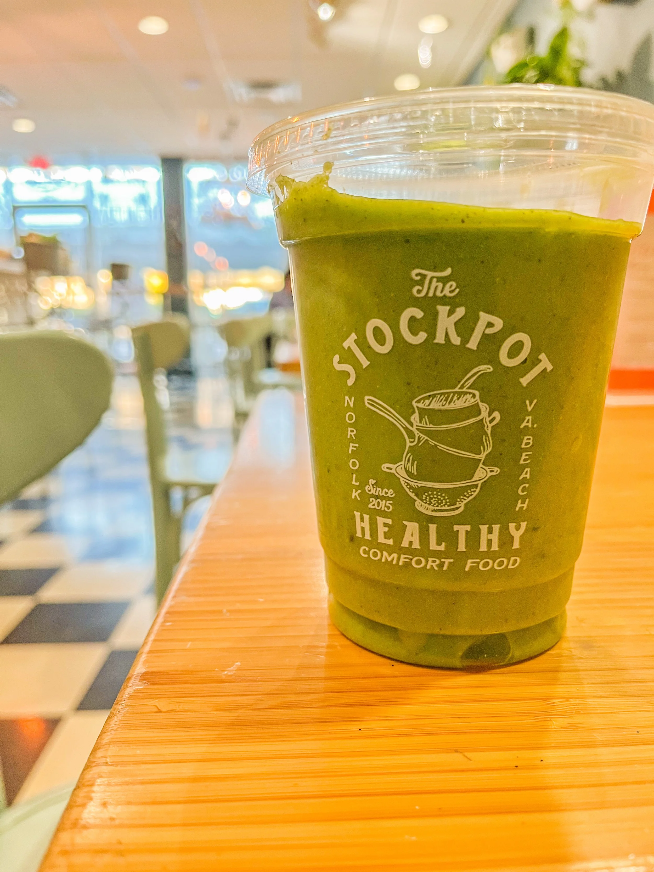 Green smoothie in a clear plastic cup with a lid, on a wooden counter at a cafe, with blurred interior background.