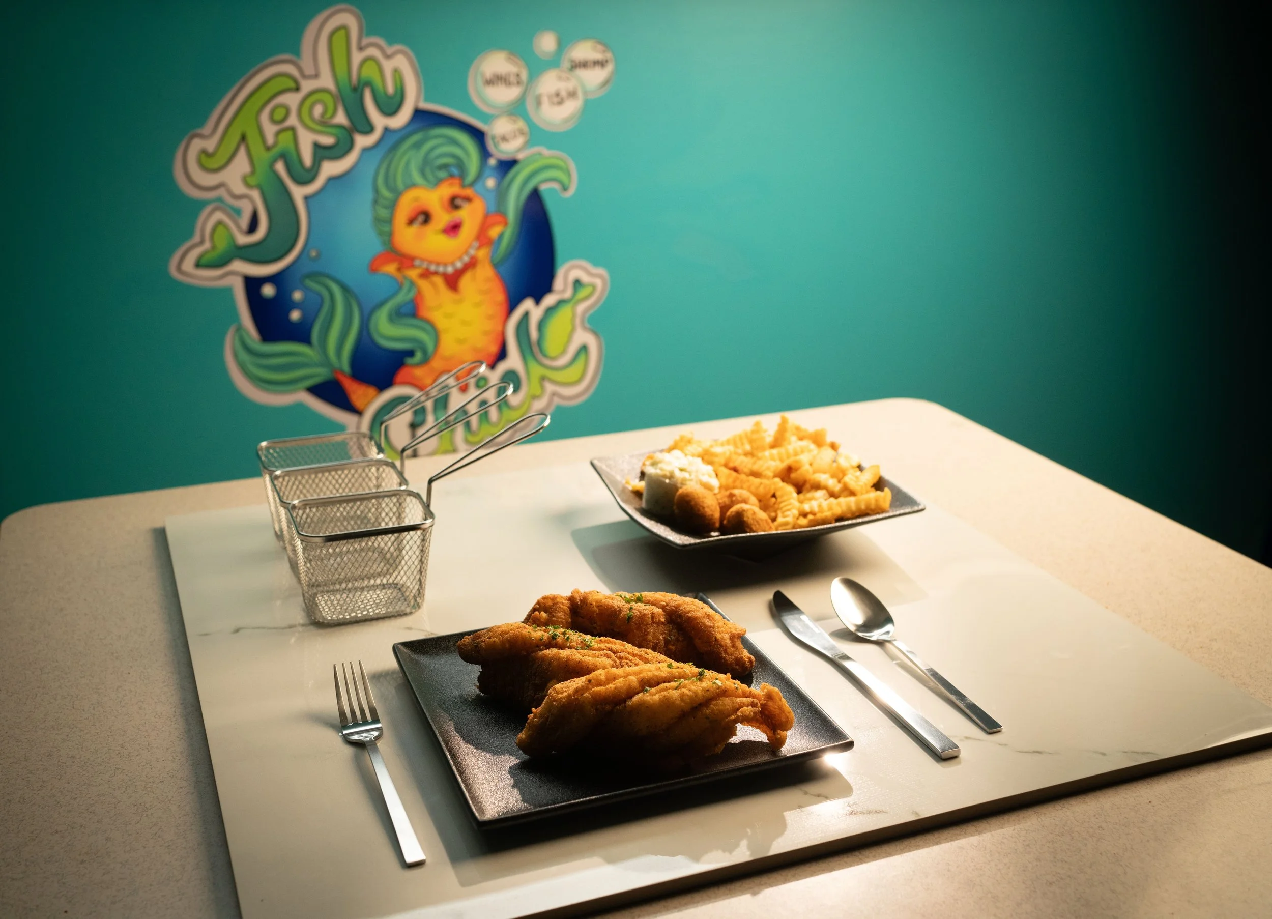 Plate of fried chicken wings on a black square plate, and a bowl of pasta with fried food on a white table, with a colorful fish-themed mural in the background.