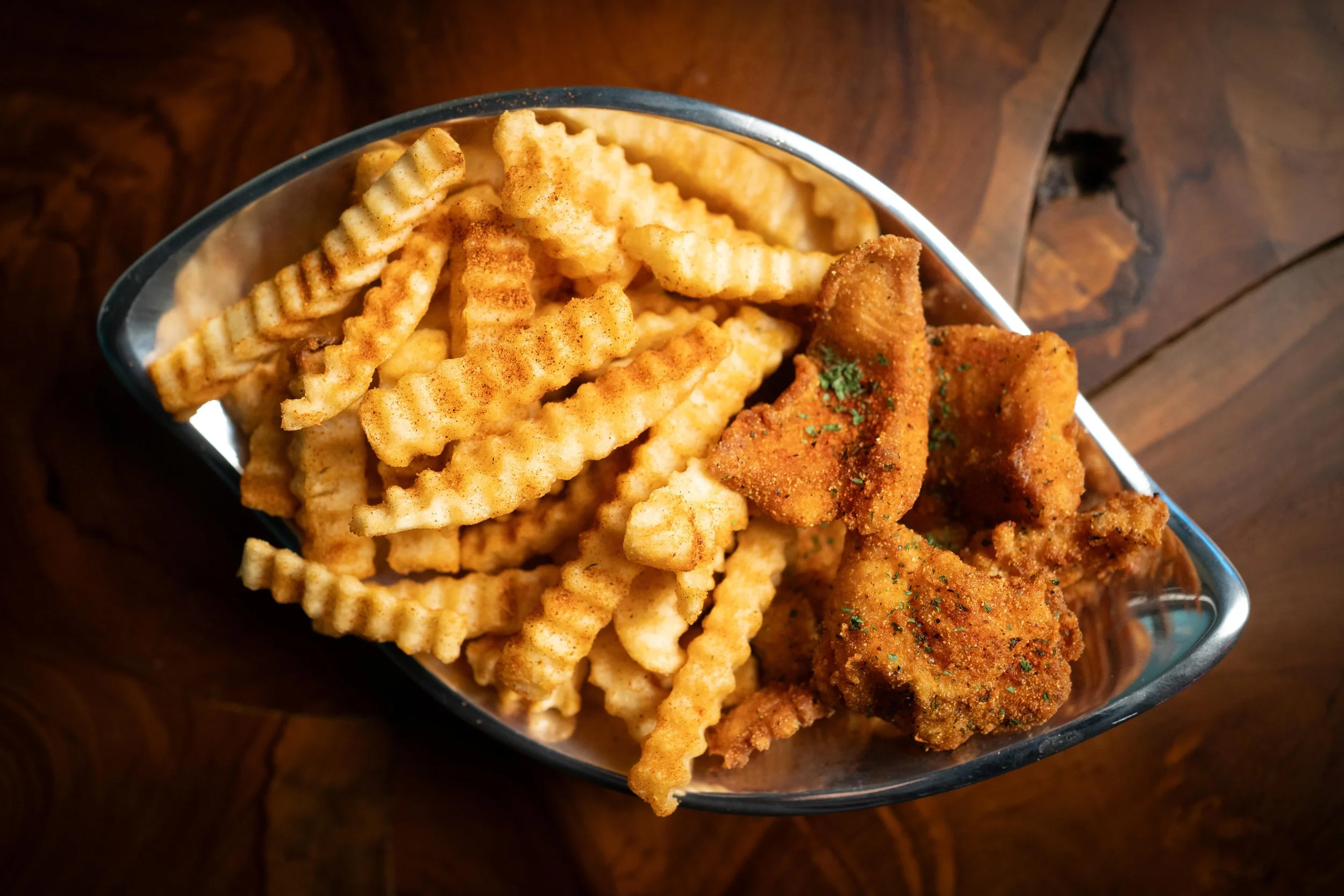 A metal dish filled with crinkle-cut French fries and fried chicken pieces, garnished with herbs, placed on a wooden surface.