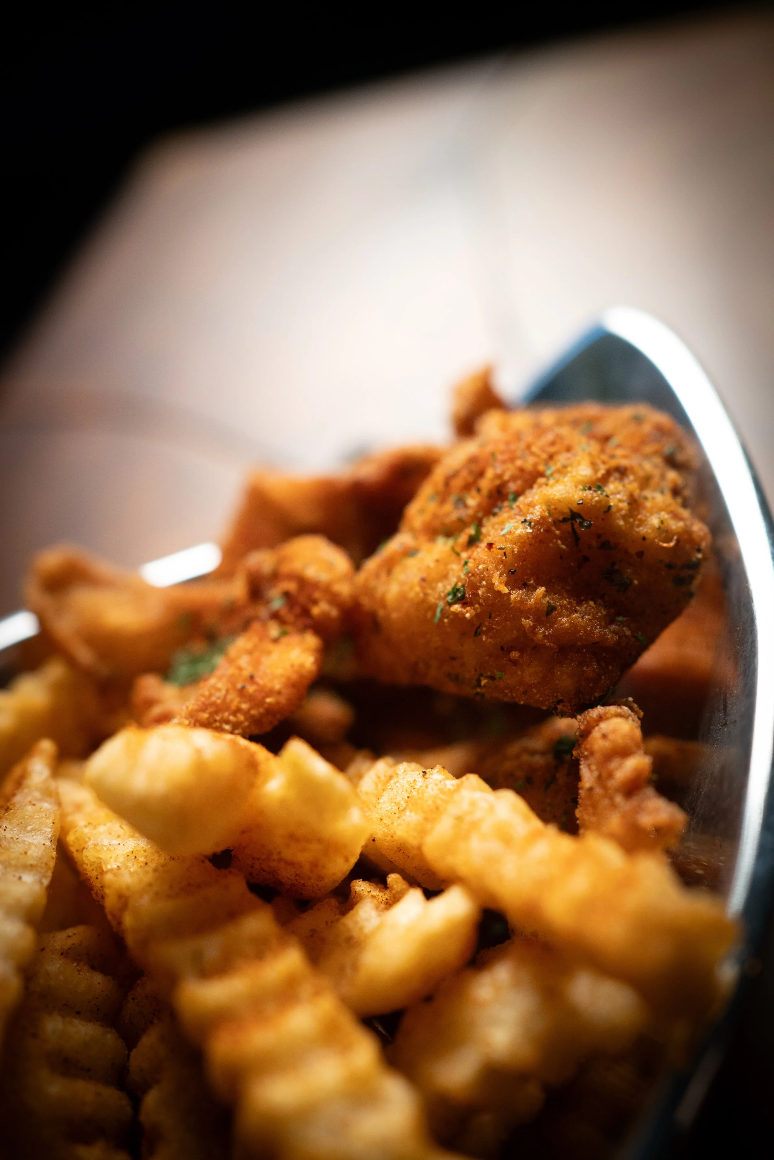 Close-up of seasoned, crispy fried chicken piece on top of crinkle-cut French fries in a metallic container.