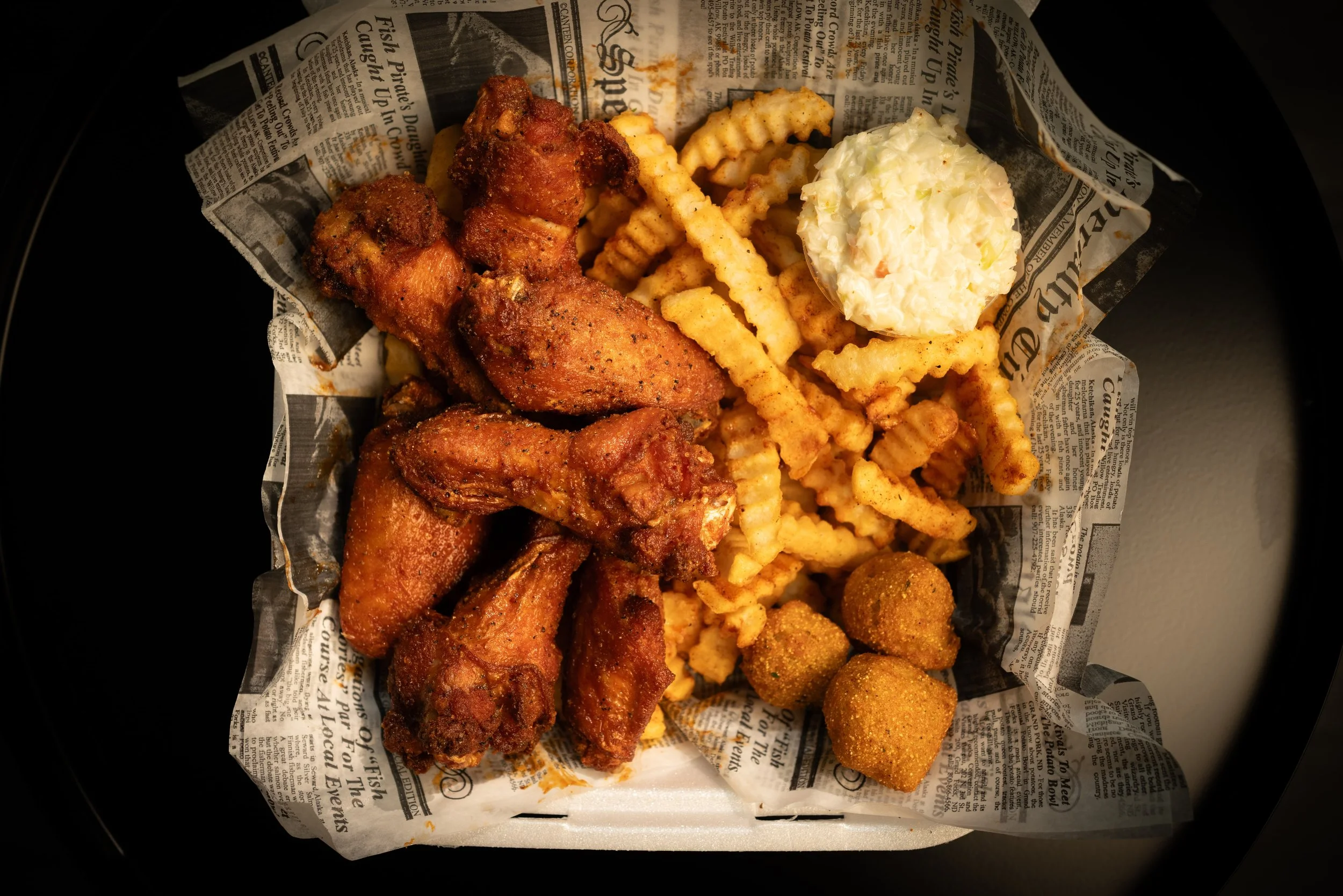 A basket of fried chicken wings, crinkle-cut French fries, a scoop of potato salad, and three hush puppies on newspaper-style paper.
