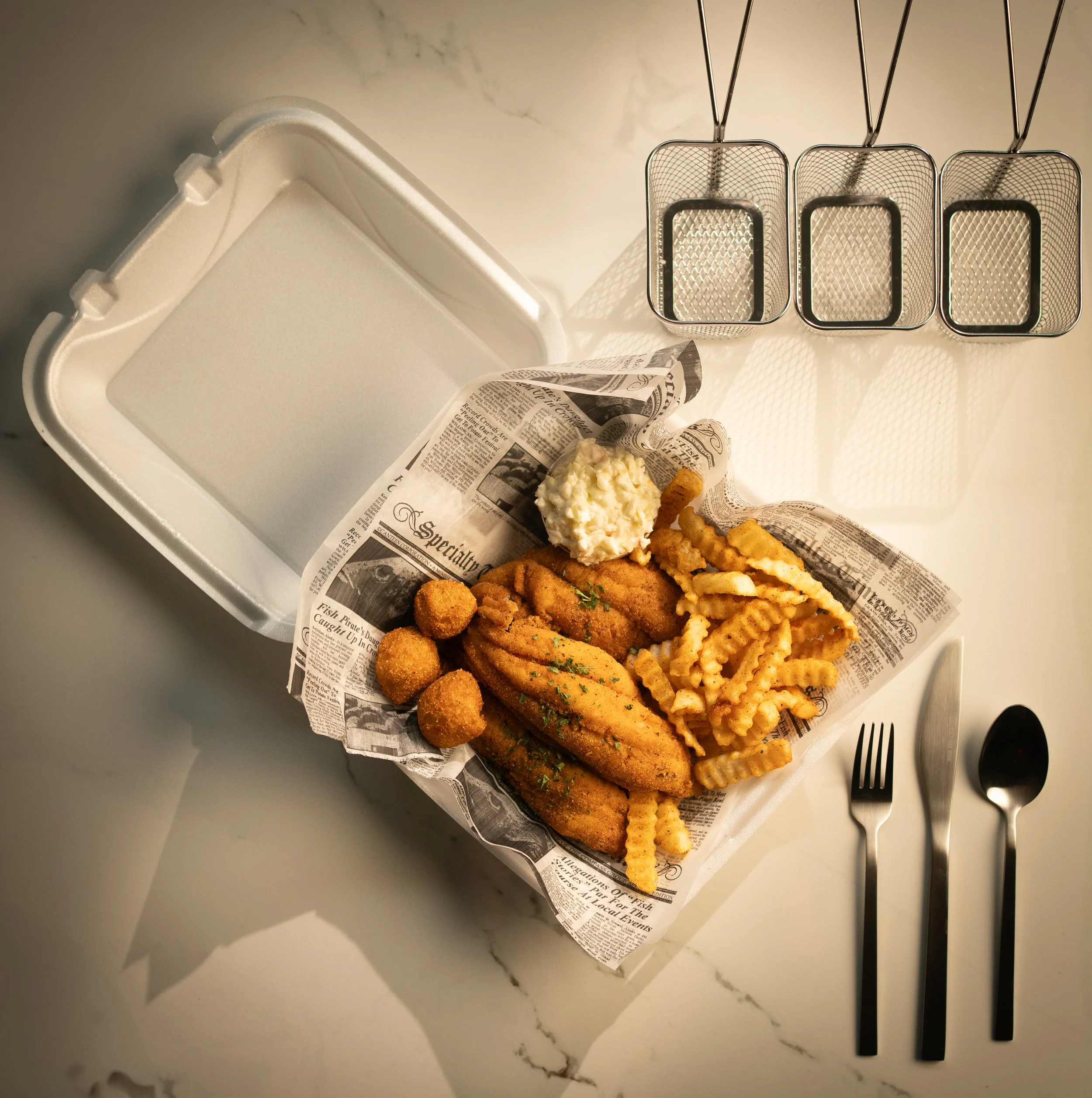 Open white takeout container with fried fish fillet, crinkle-cut fries, hush puppies, and a scoop of coleslaw, lined with newspaper print paper, on a white marble surface with a fork, knife, and spoon beside it.
