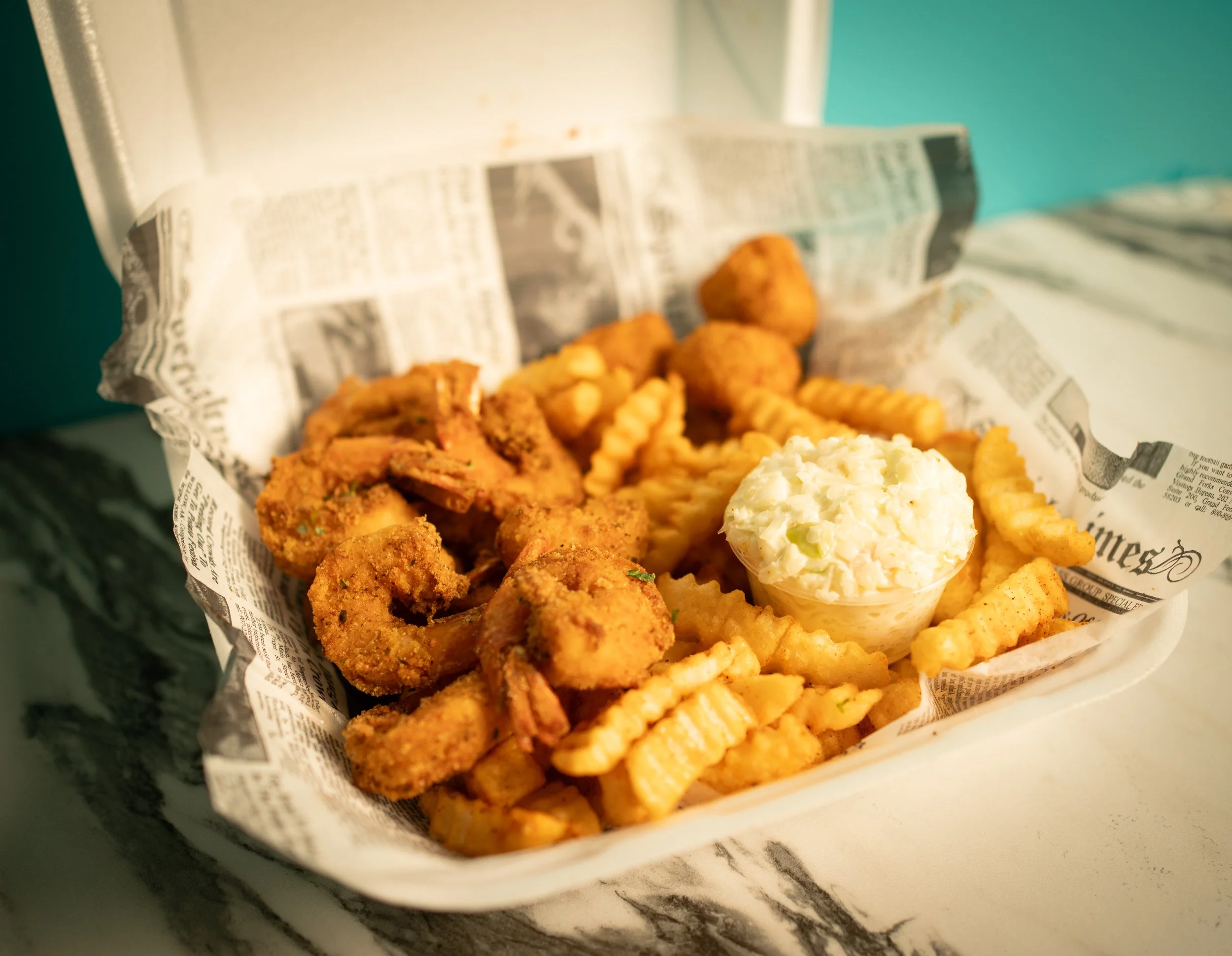 Fried shrimp, crinkle-cut fries, coleslaw in a paper food tray lined with newspaper-style paper.