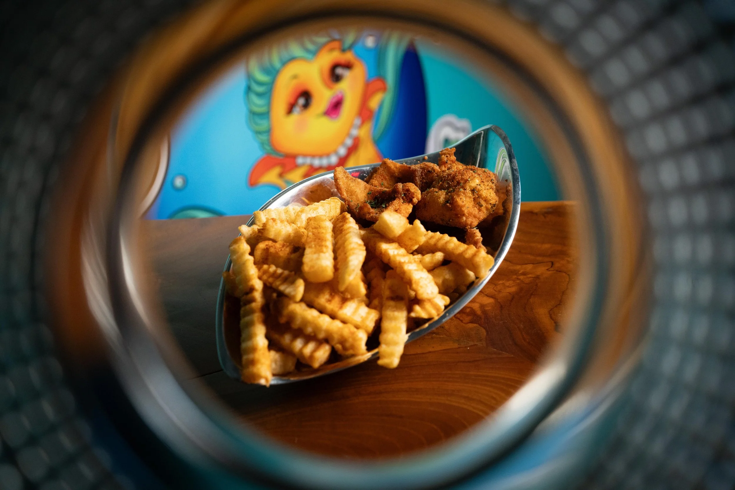 View of fried chicken and French fries on a tray, seen through the opening of a metal food container, with a colorful cartoon fish mural in the background.