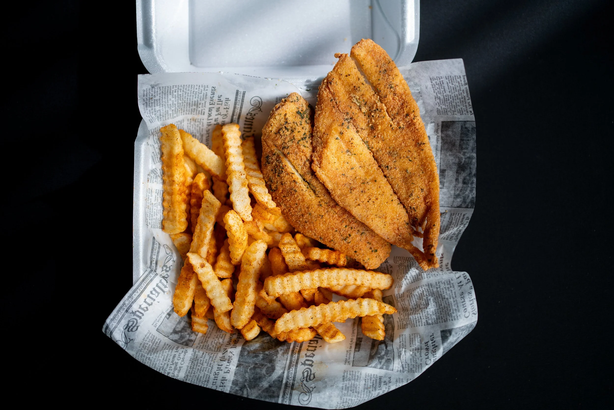 Takeout box with fried fish fillets and crinkle-cut French fries on black background.