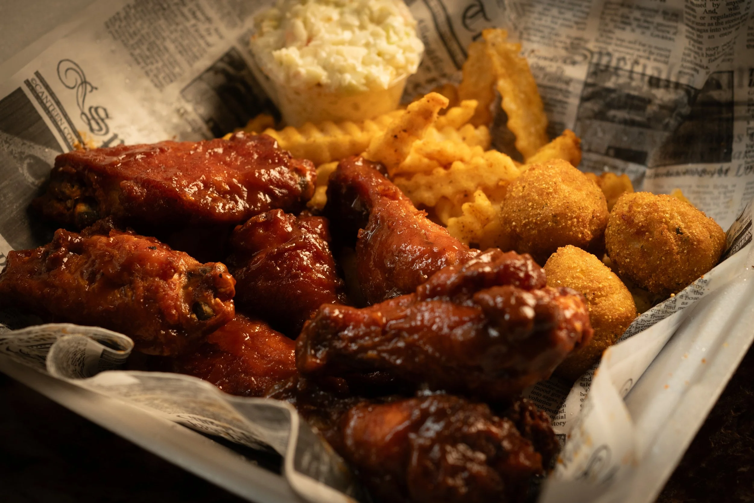 Basket of fried chicken wings, breaded hush puppies, waffle fries, and a cup of potato salad on newspaper-lined tray.