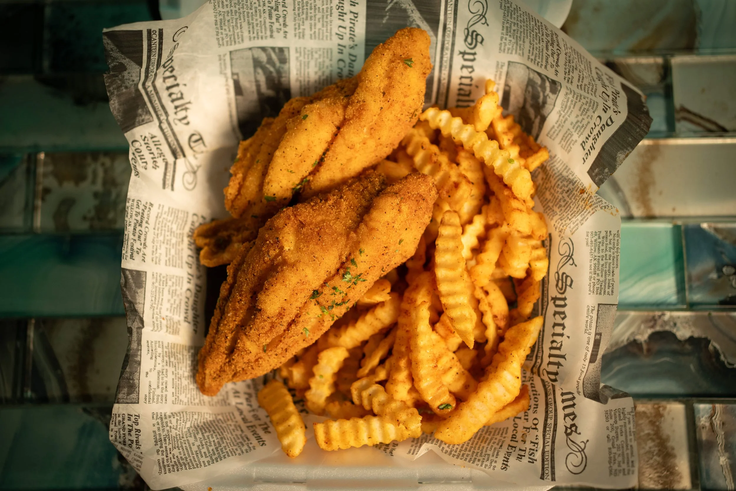 Basket of fried fish fillets and crinkle-cut French fries on newspaper-style paper.