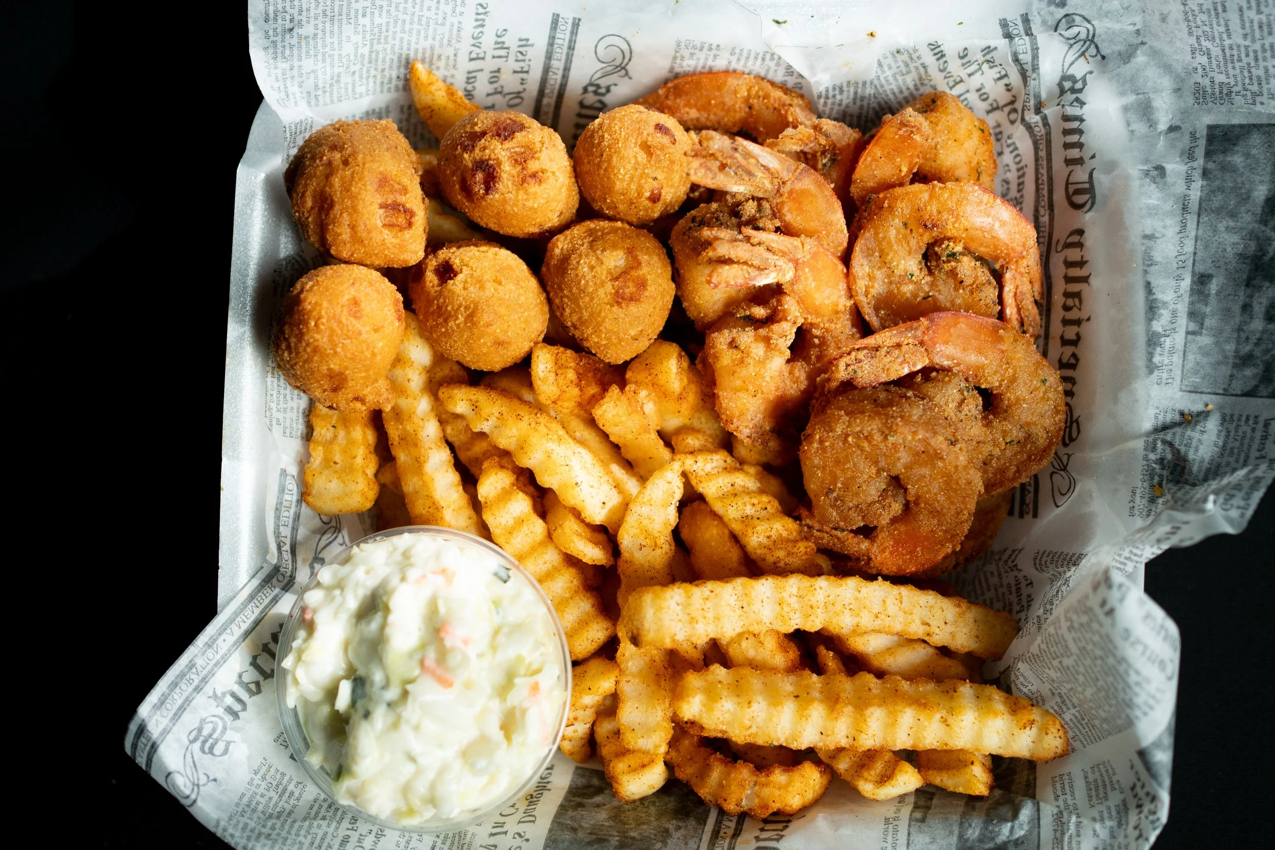A basket of fried seafood including shrimp, fish, crab balls, and French fries with a side of coleslaw on newspaper-lined paper.