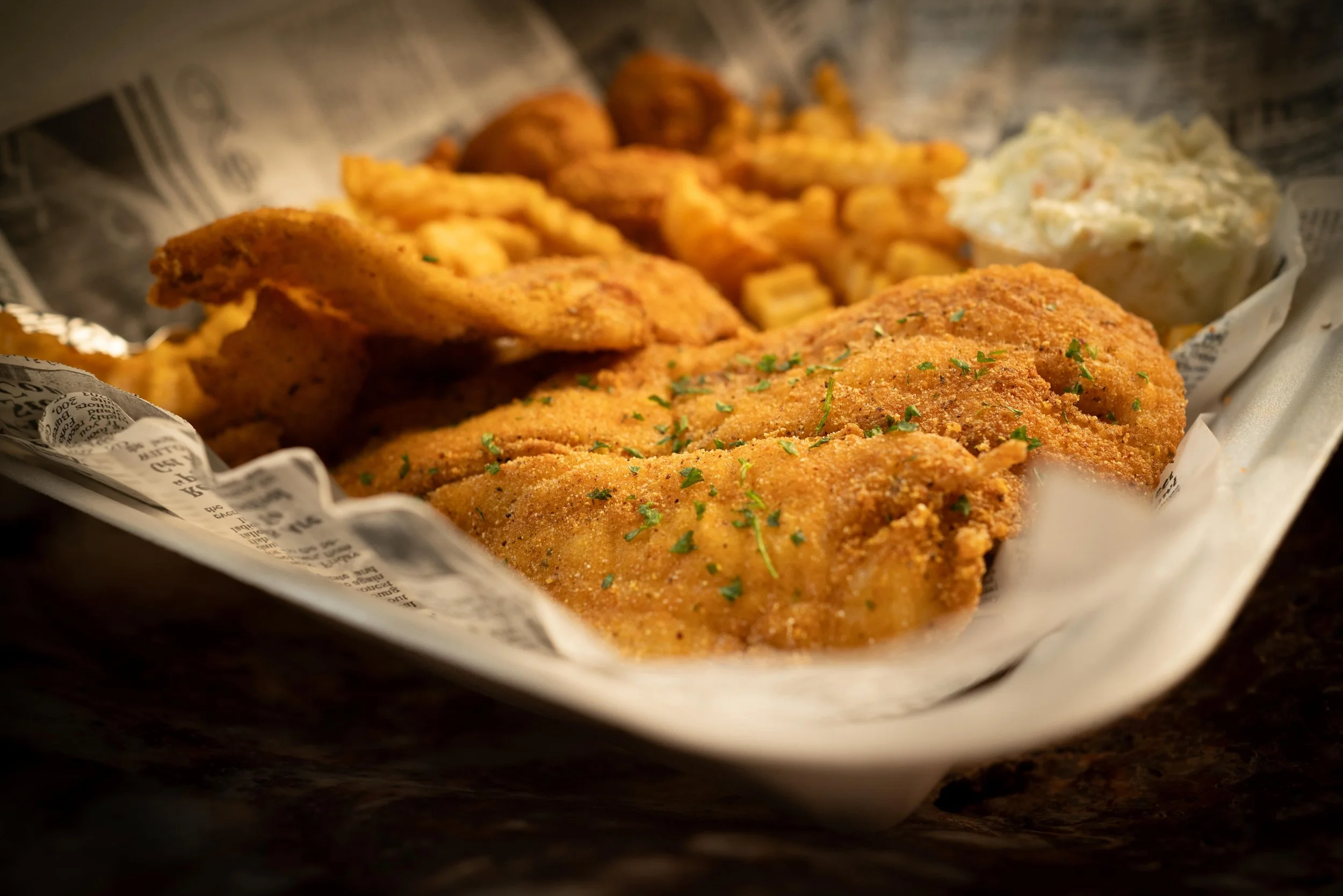 Fried fish fillet with a sprinkle of parsley, served with coleslaw and fried potato wedges on a newspaper-lined tray.