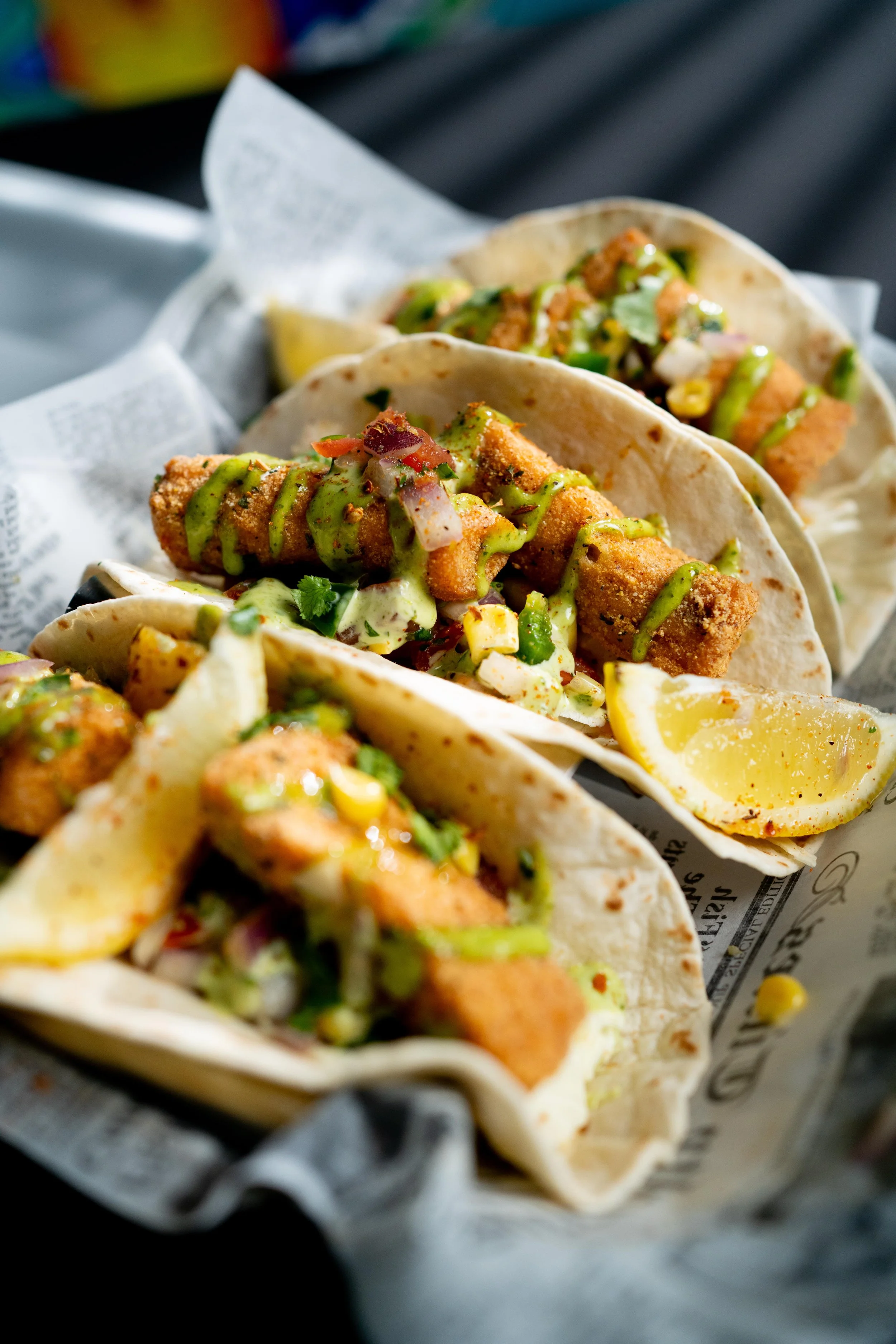 Three fish tacos with fried fish fillets, topped with chopped onions, cilantro, and drizzled with green sauce, accompanied by lemon wedges, on a tray lined with newspaper.