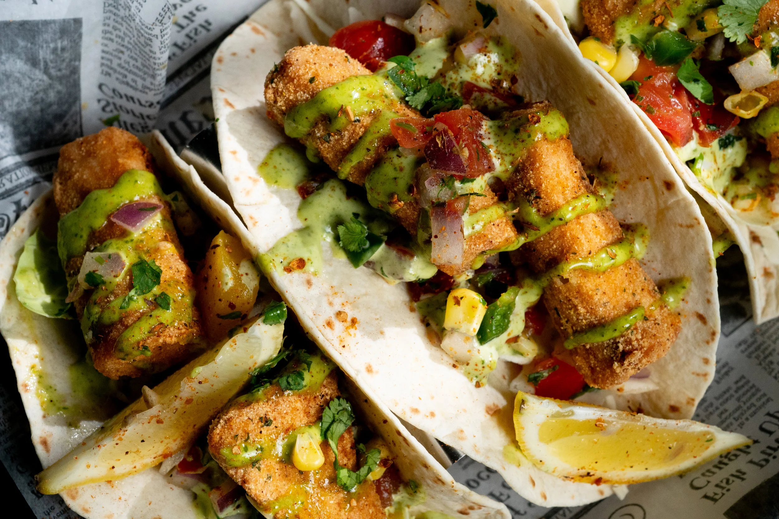 Three fish tacos topped with avocado sauce, chopped tomatoes, onions, and cilantro, served with lemon wedges on a newspaper-lined tray.