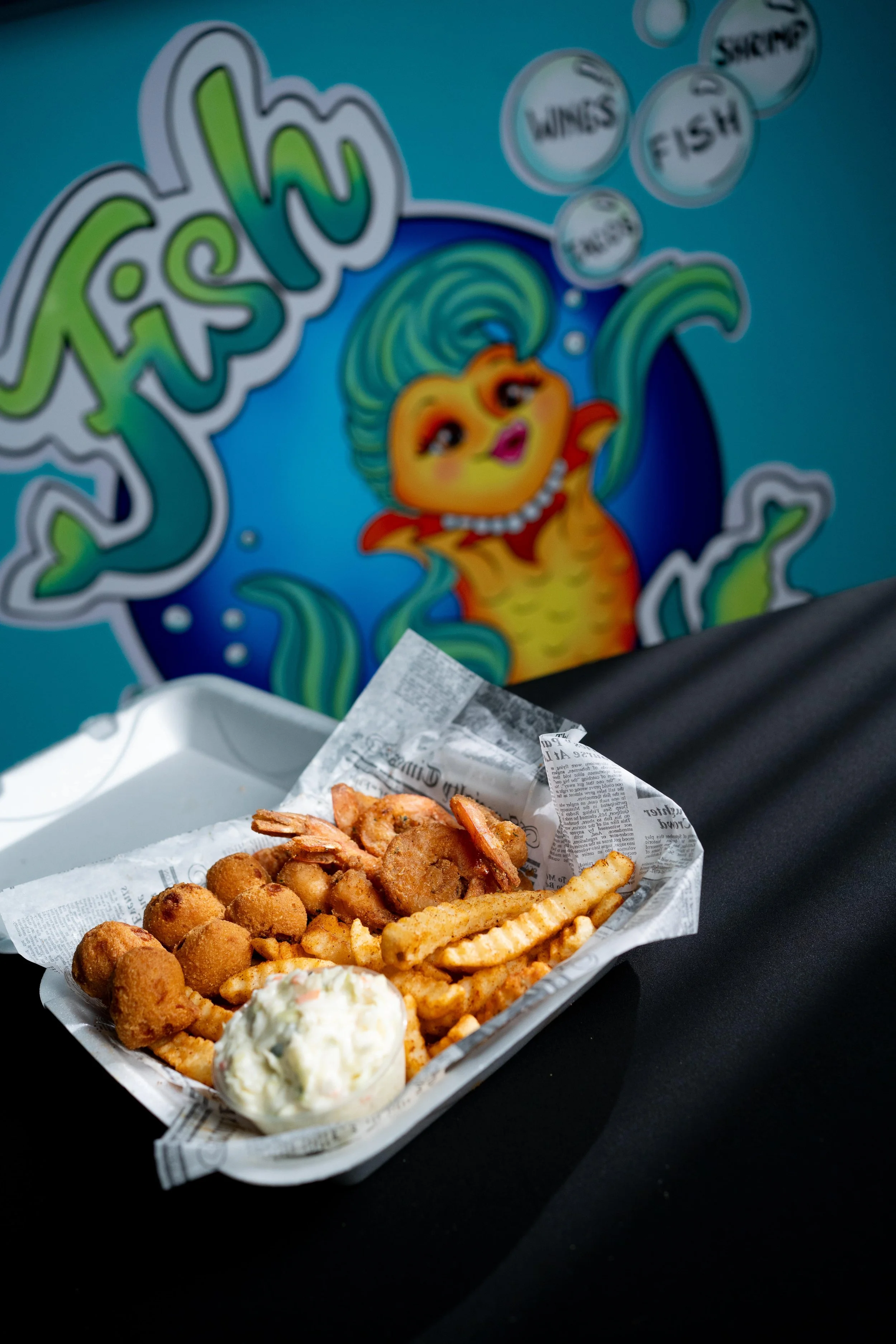 A tray of fried seafood and French fries with a dollop of tartar sauce in front of a colorful mural with a fish character and the word "fish" in bold letters.