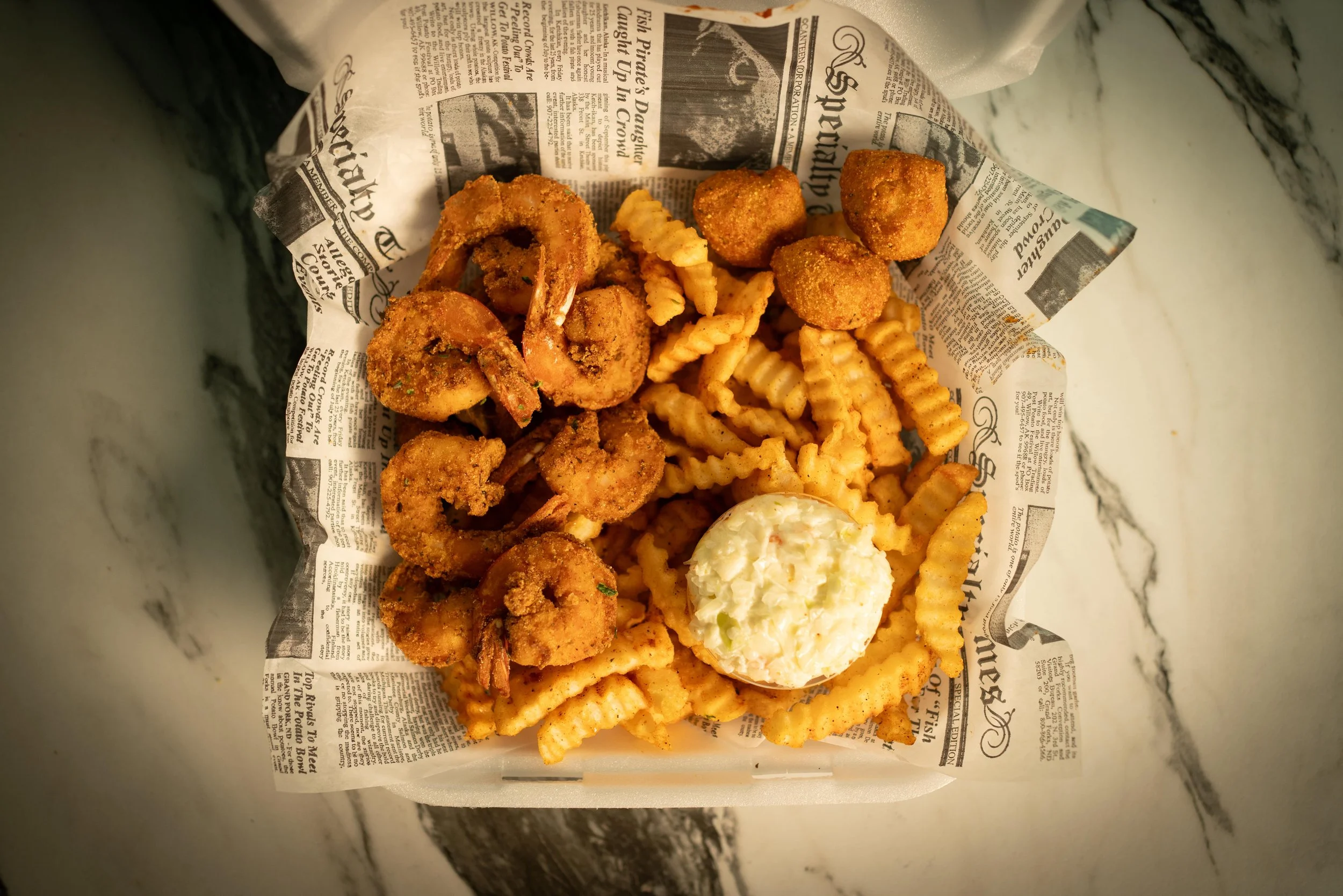 Basket of fried shrimp, crinkle-cut French fries, and a scoop of mayonnaise or tartar sauce on newspaper-style parchment paper, on a white marble surface.