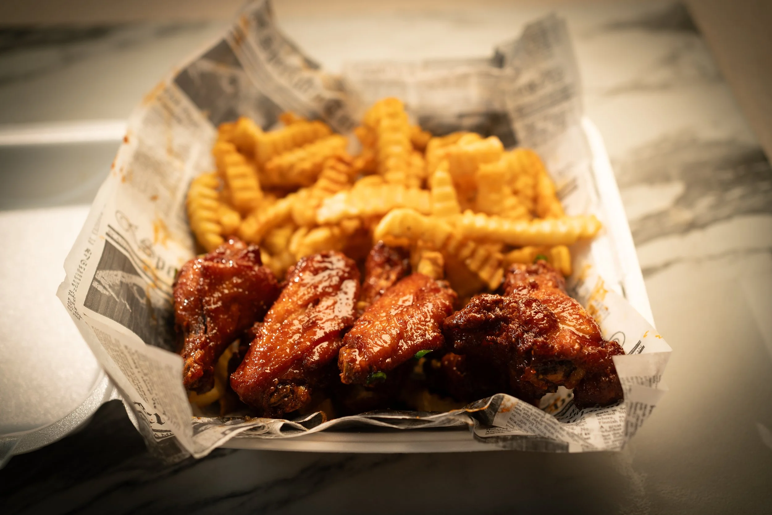 Basket of crispy French fries and glazed chicken wings on newspaper-lined container