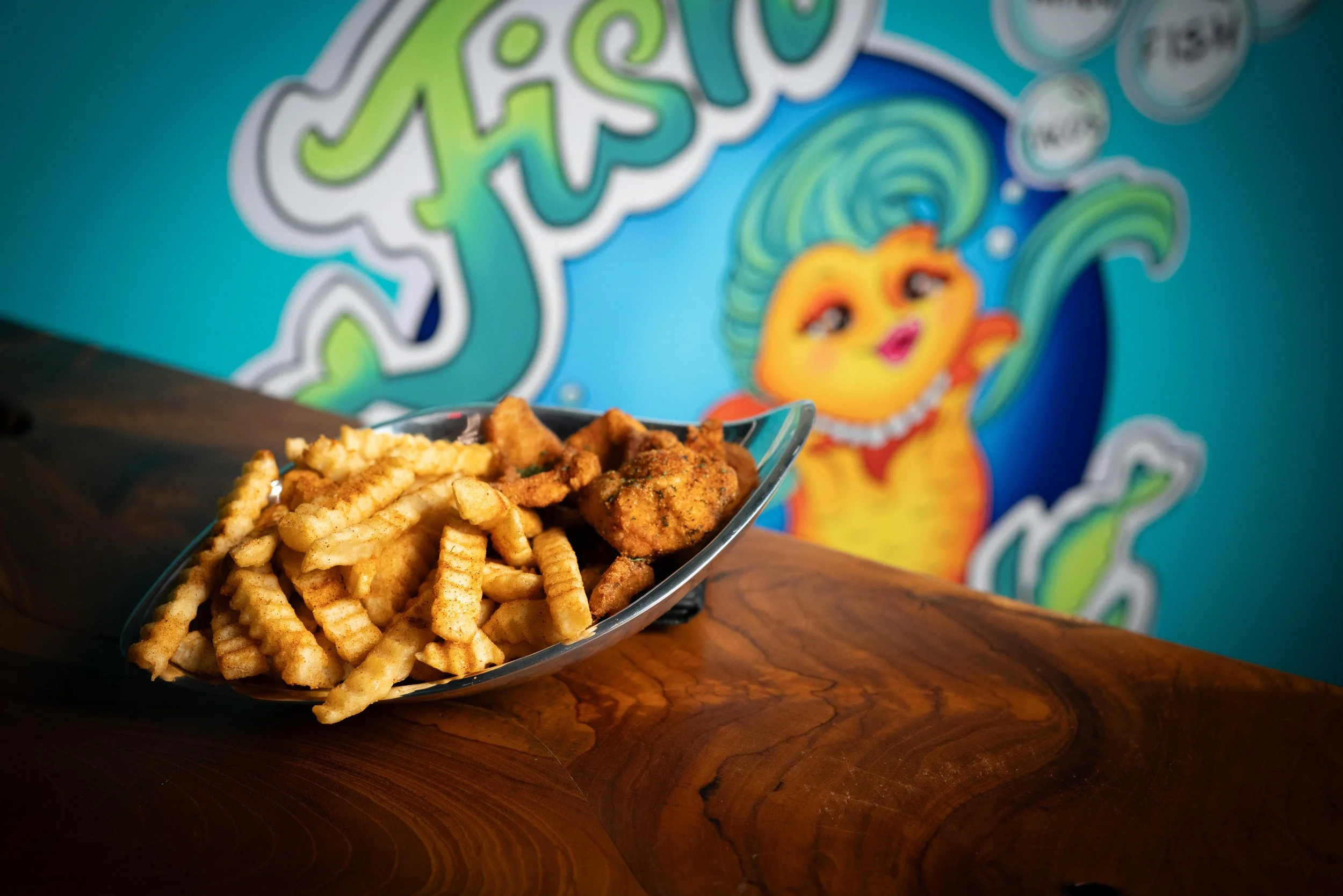 A metal tray filled with French fries and fried chicken on a wooden table with a colorful cartoon mural of a turquoise mermaid in the background.