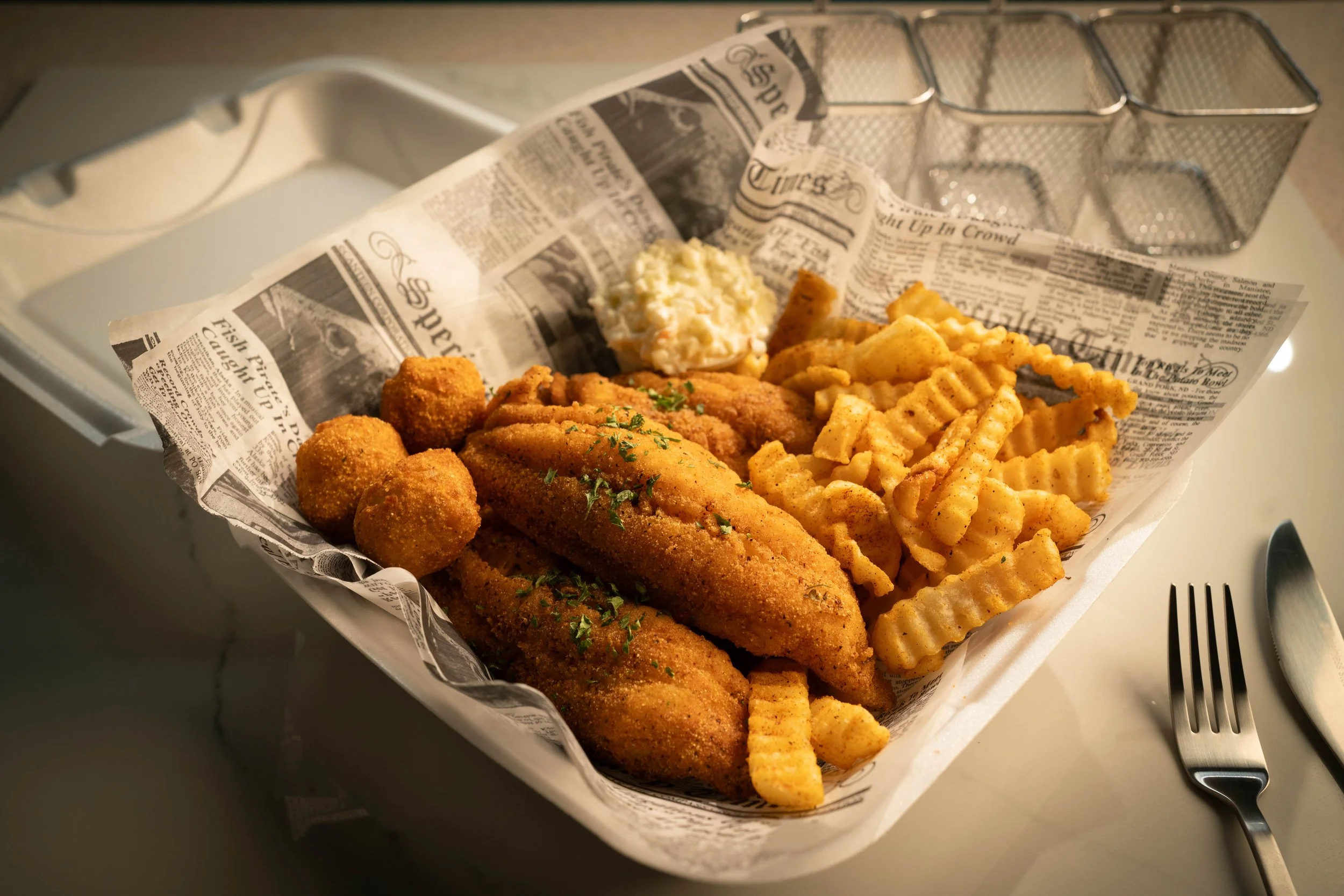 Basket of fried fish fillets, crinkle-cut French fries, hush puppies, and coleslaw on newspaper-lined tray with fork and knife nearby.