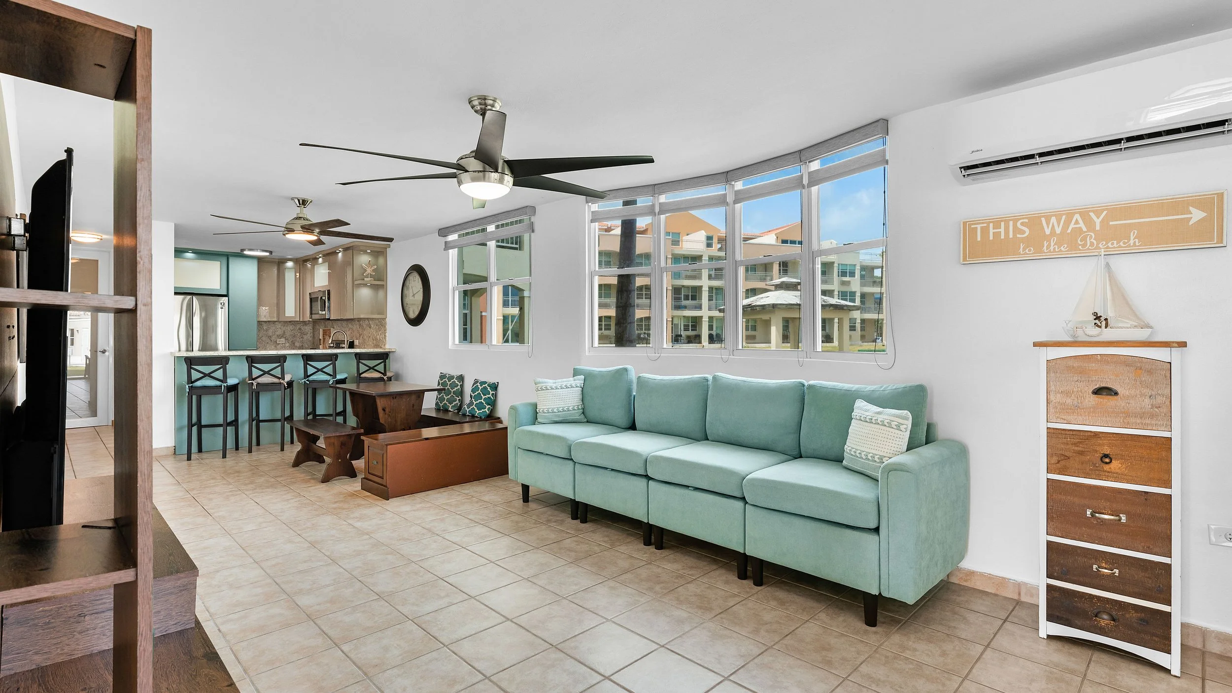 Living room with a light green sofa, tiled floor, white walls, large windows showing apartment buildings outside, and a corner sign that says "This way to the beach." There is a wooden storage drawer and nautical decor in Puerto Rico.