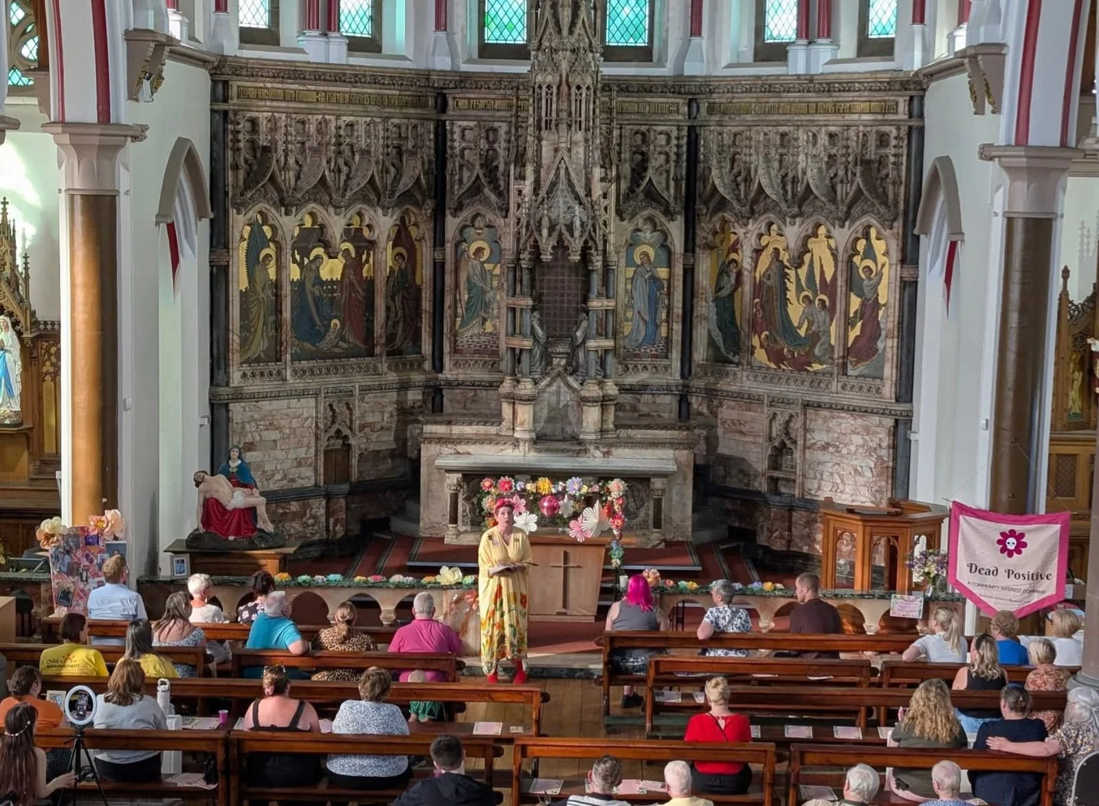 A church interior with a woman (Kirstie) speaking at a lectern decorated with flowers, an audience seated on wooden pews, and religious art and statues behind the speaker.