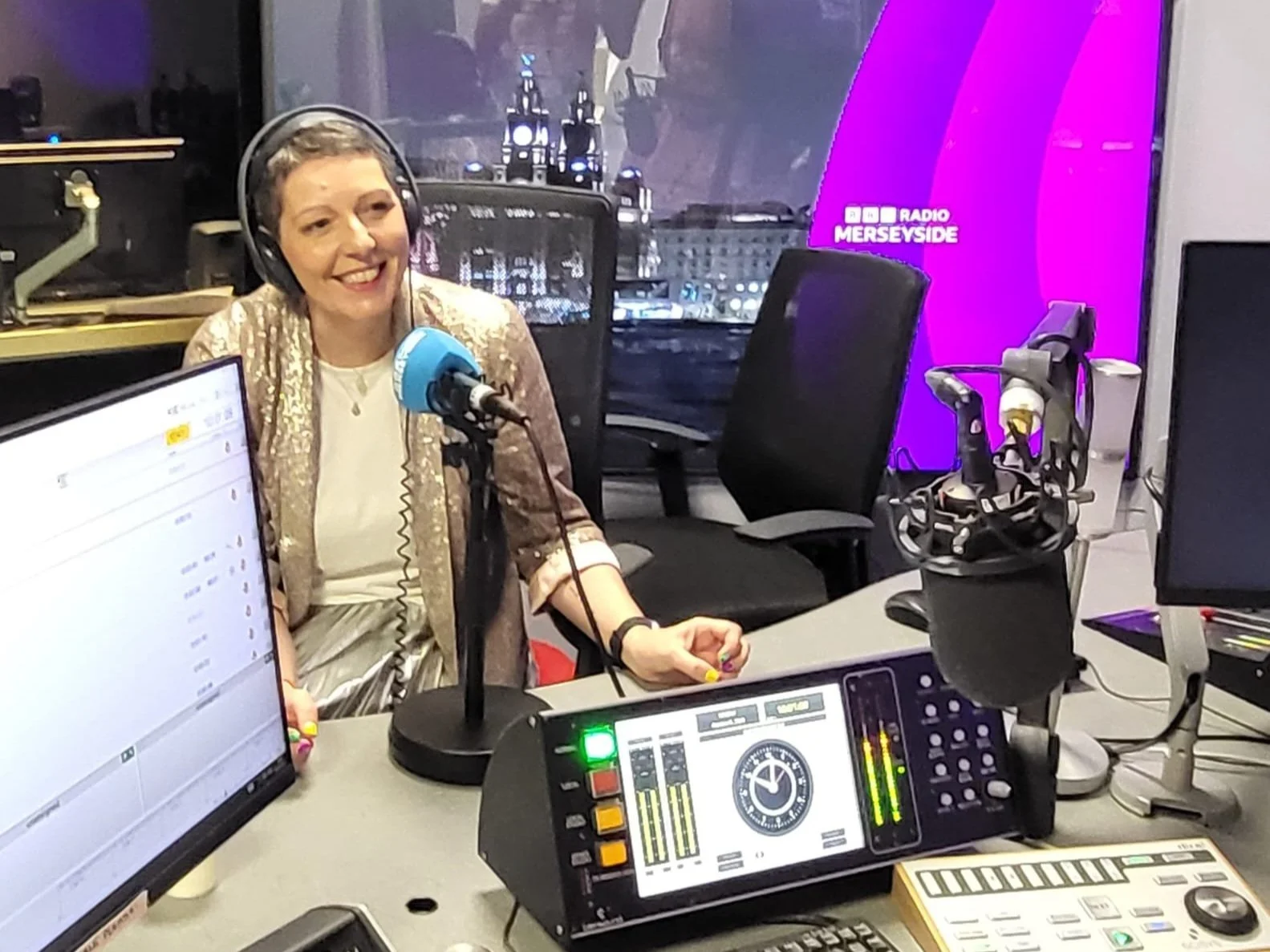 A woman, Kirstie, with short gray hair, wearing a pearl-colored blazer, smiling while speaking into a blue microphone in a BBC Radio studio with broadcasting equipment and computer screens.