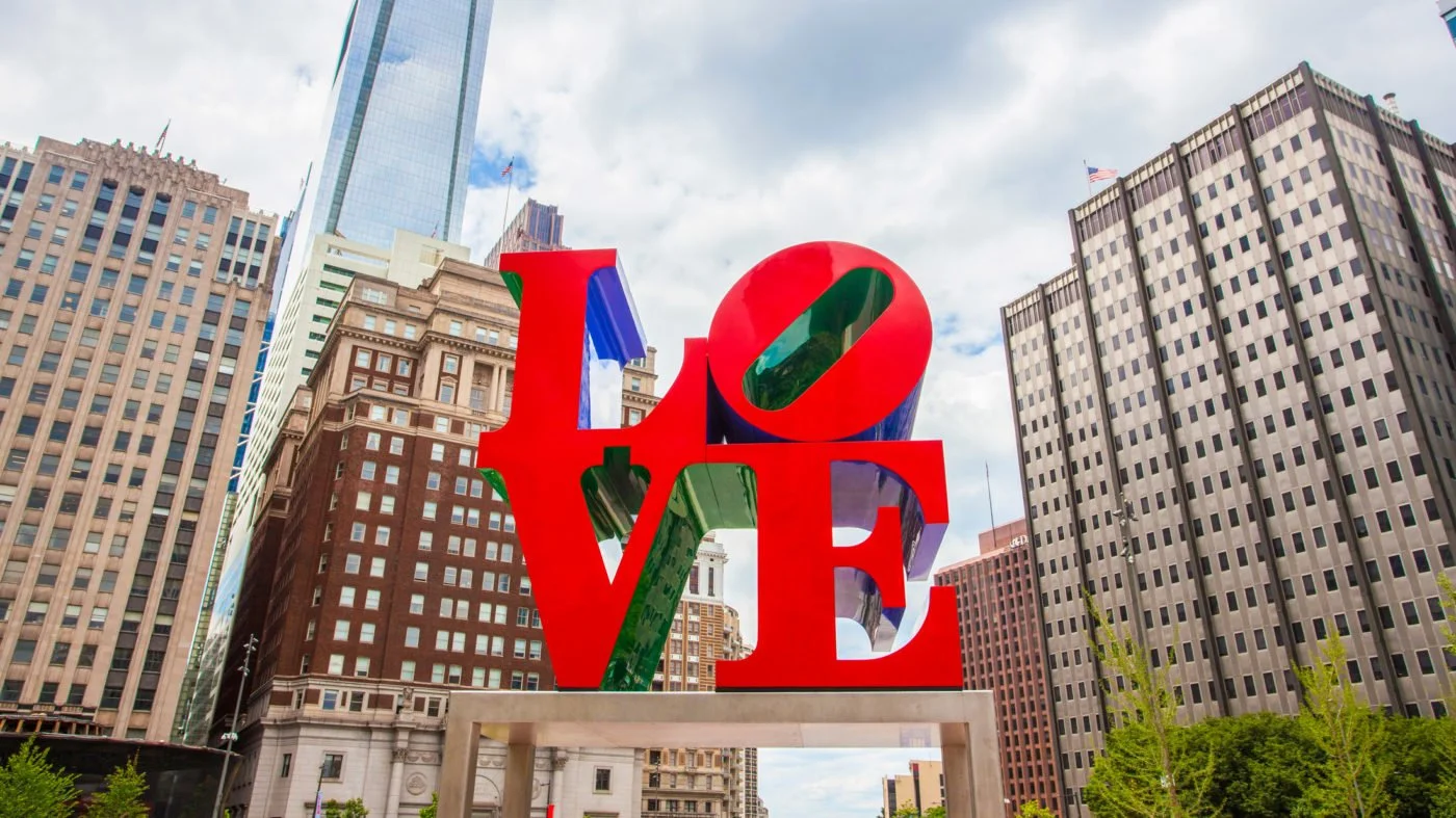 Large colorful sculpture of the word 'LOVE' with the 'O' tilted, set against a backdrop of tall city buildings and a partly cloudy sky in an urban park.
