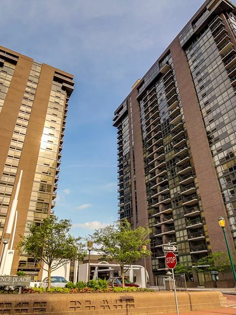 Two tall modern residential buildings with glass balconies, trees, and a street view in an urban setting.