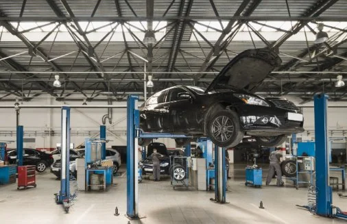A black sedan car is lifted on an automotive lift in a spacious car repair shop, with mechanics working underneath and other vehicles in the background.