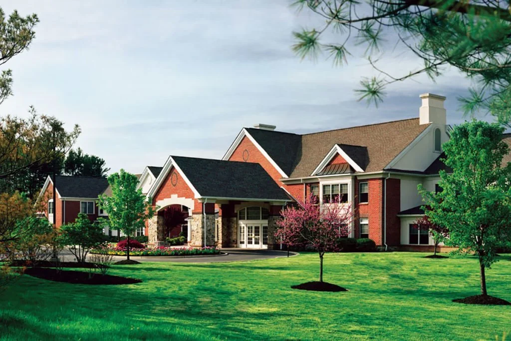 A large brick house with multiple gables and a covered entrance, surrounded by a green lawn and various trees, some flowering with pink blossoms.