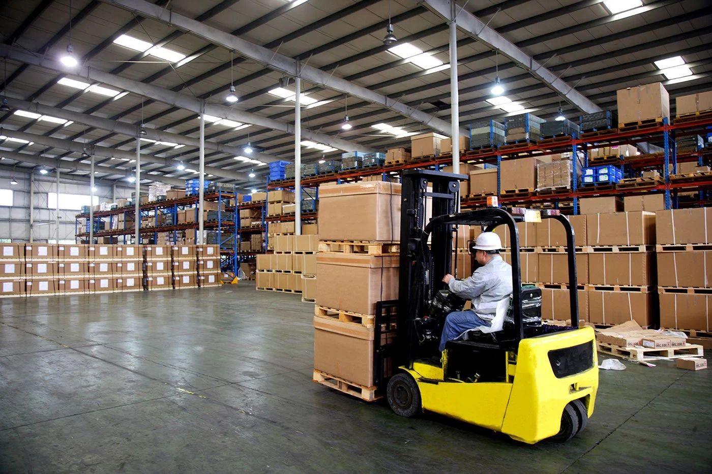 A worker in a warehouse operating a yellow forklift with stacks of cardboard boxes and shelves filled with boxes in the background.