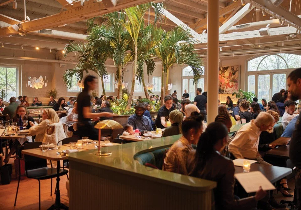 A bustling restaurant with many patrons seated at tables, tropical palm trees in the center, and natural light coming through large windows.