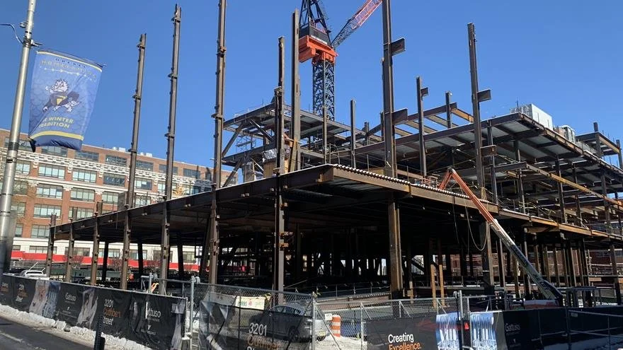 Construction site with steel framework and crane, in an urban area with buildings and a winter-themed banner nearby.