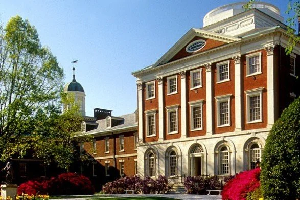 A historic university building with red brick and white columns, surrounded by greenery and colorful flowers.